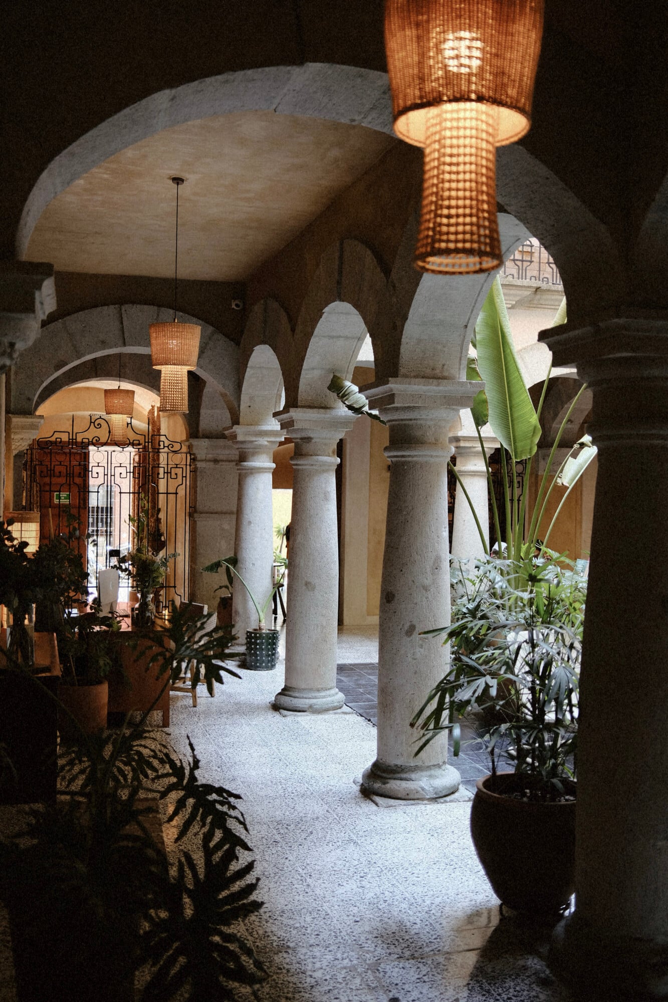 Casa Antonieta, Mexico — Colonnade with cylindrical columns, arched ceilings, woven pendant lights, potted plants, and stone flooring in a Mediterranean-style courtyard.
