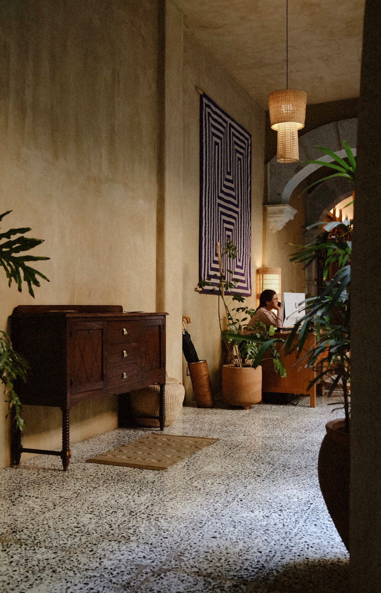 Casa Antonieta, Mexico — Concrete corridor with woven pendant light, dark wood cabinet, potted plants, and geometric textile wall hanging casting warm shadows.