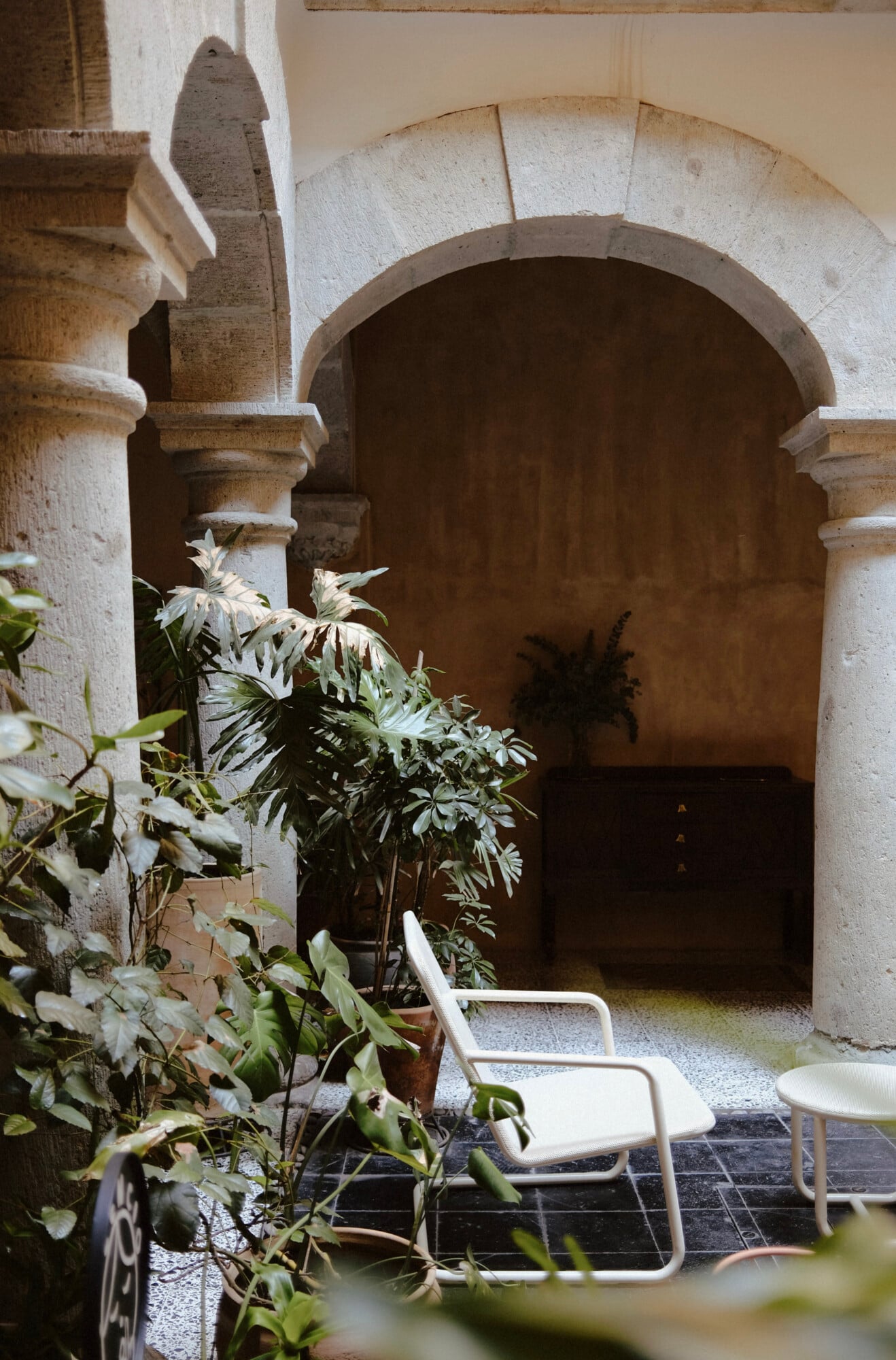Casa Antonieta, Mexico — Stone archway with wooden door, potted flowers, and modernist white chair in sunlit courtyard with terracotta tiles.