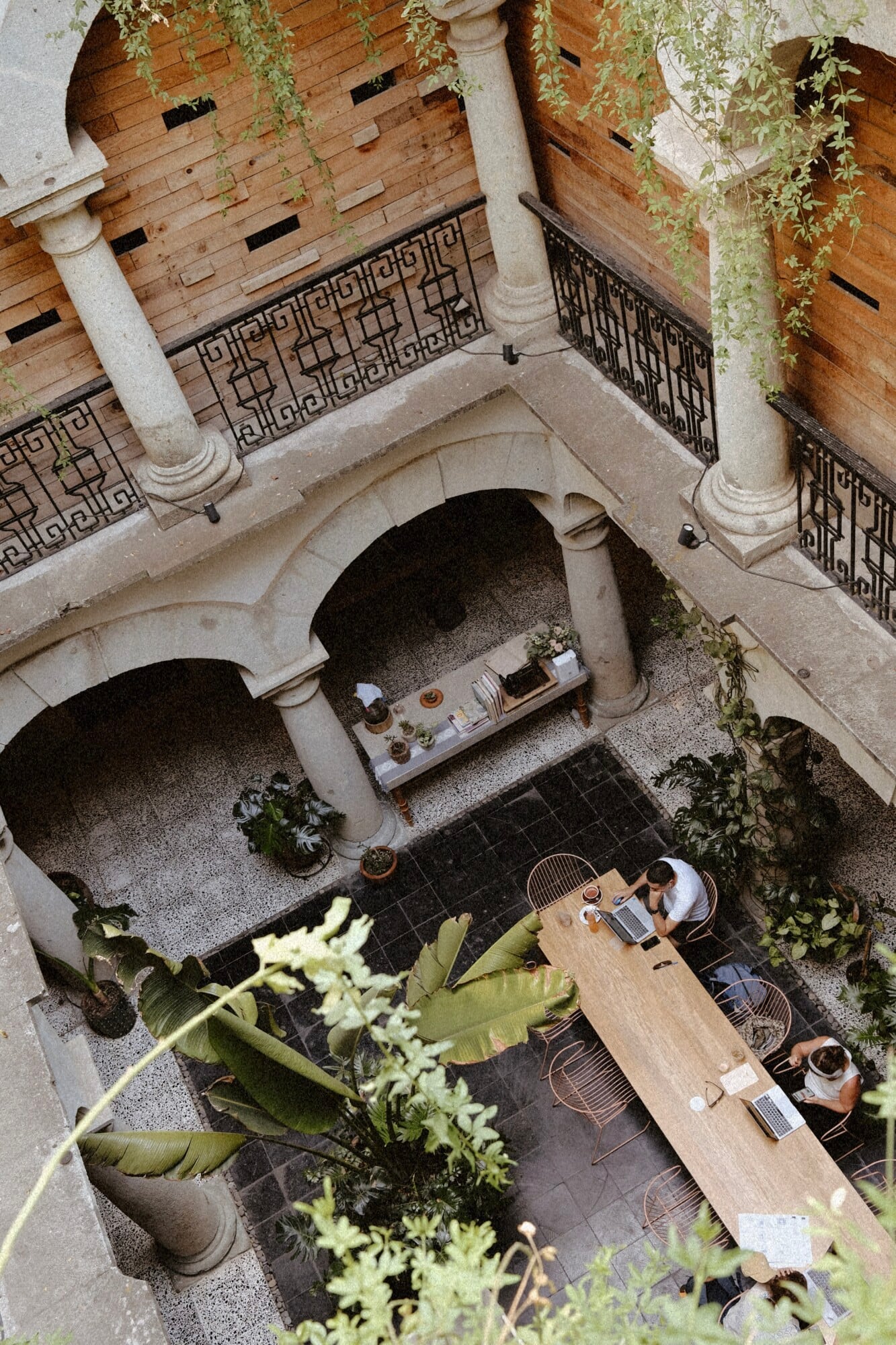 Casa Antonieta, Mexico — Overhead view of courtyard with arched colonnades, geometric ironwork railings, stone columns, cascading ivy, and guests seated at wooden table on dark tile flooring.