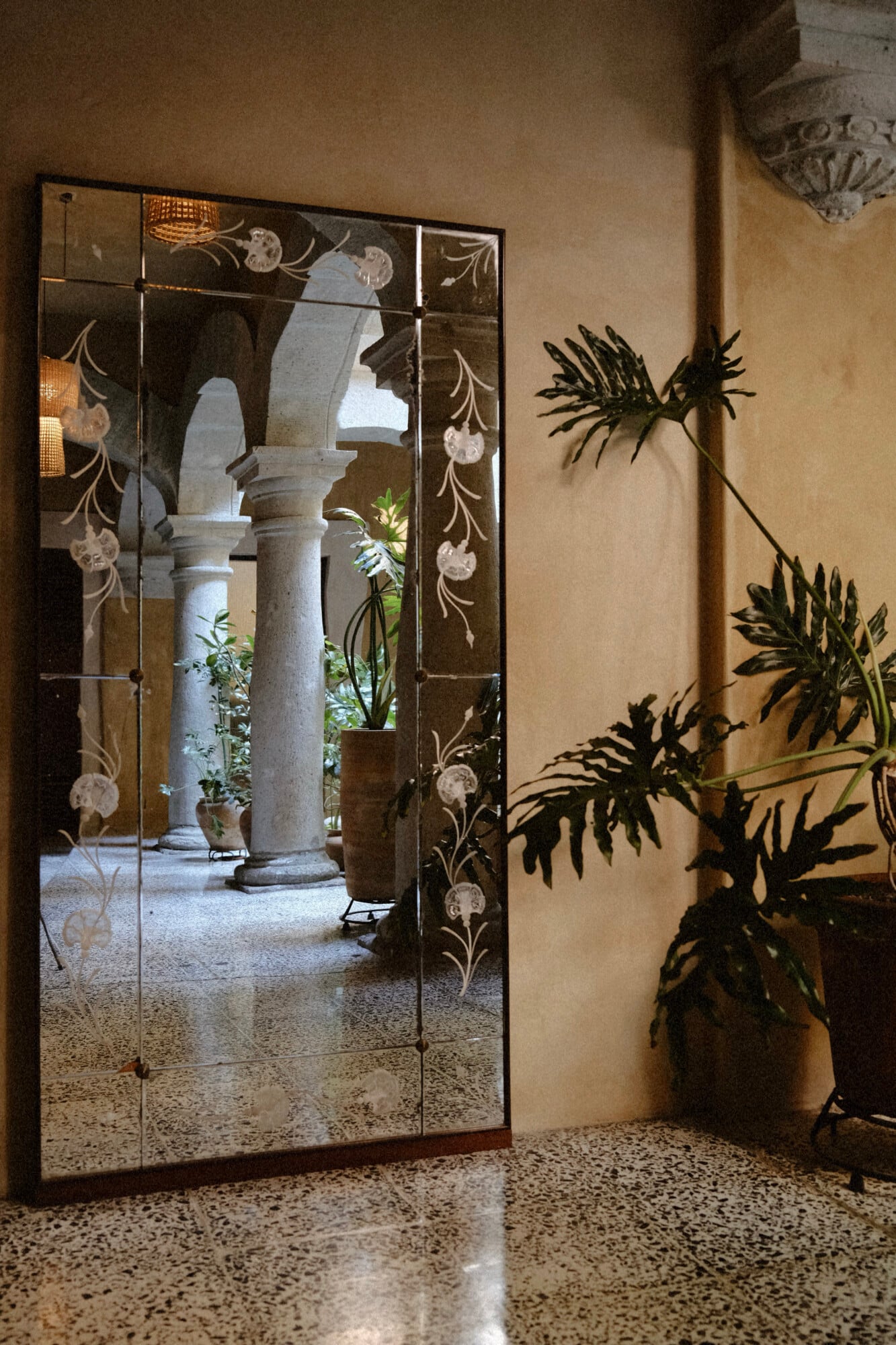 Casa Antonieta, Mexico — Decorative mirror with painted glass frame reflecting colonnaded courtyard, potted fern casting shadow on textured wall.