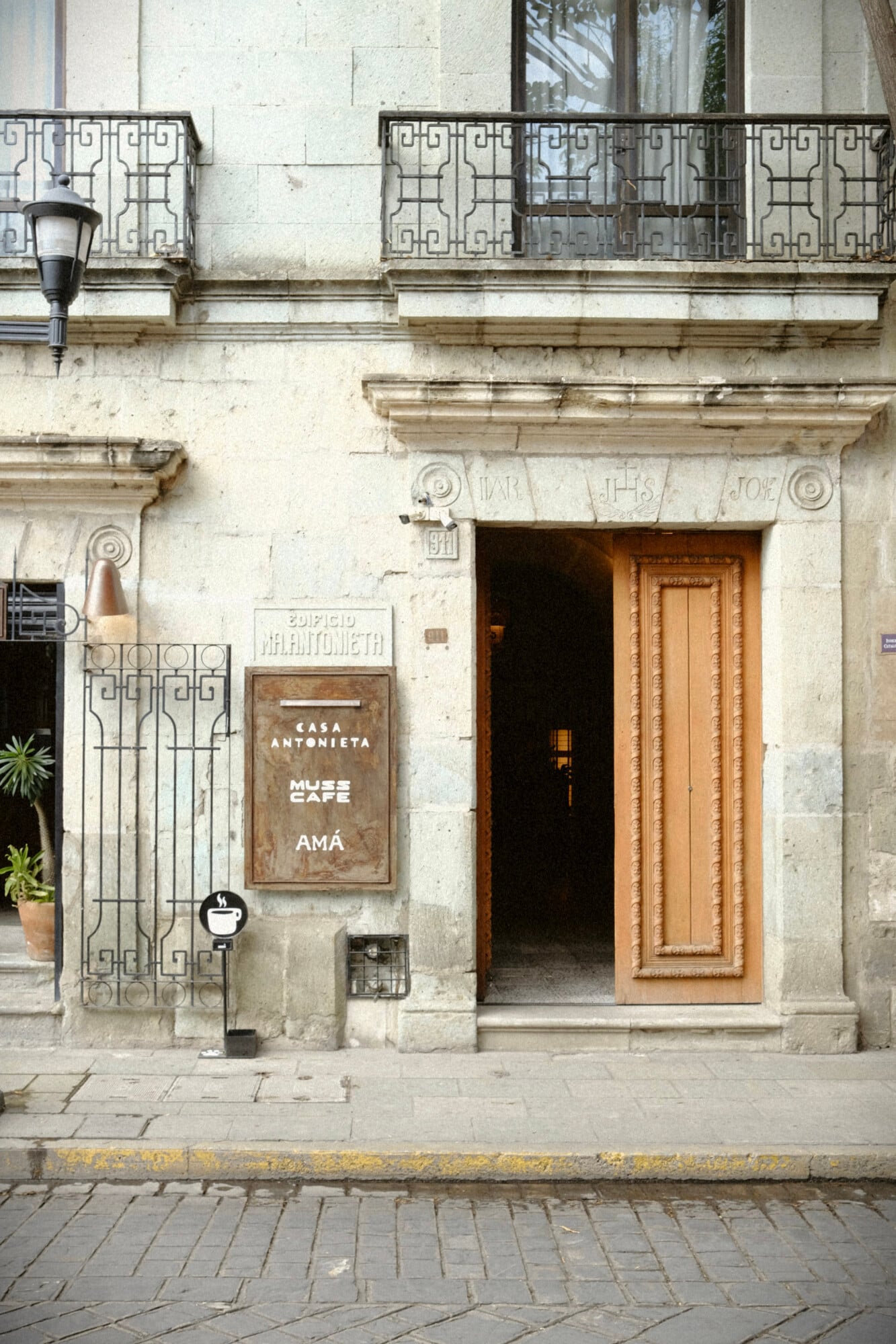 Muss Café, Mexico — Neoclassical facade with ornate wooden door, geometric ironwork balconies, stone archway, and weathered cream-colored walls displaying heritage architecture and bohemian cafe aesthetic.