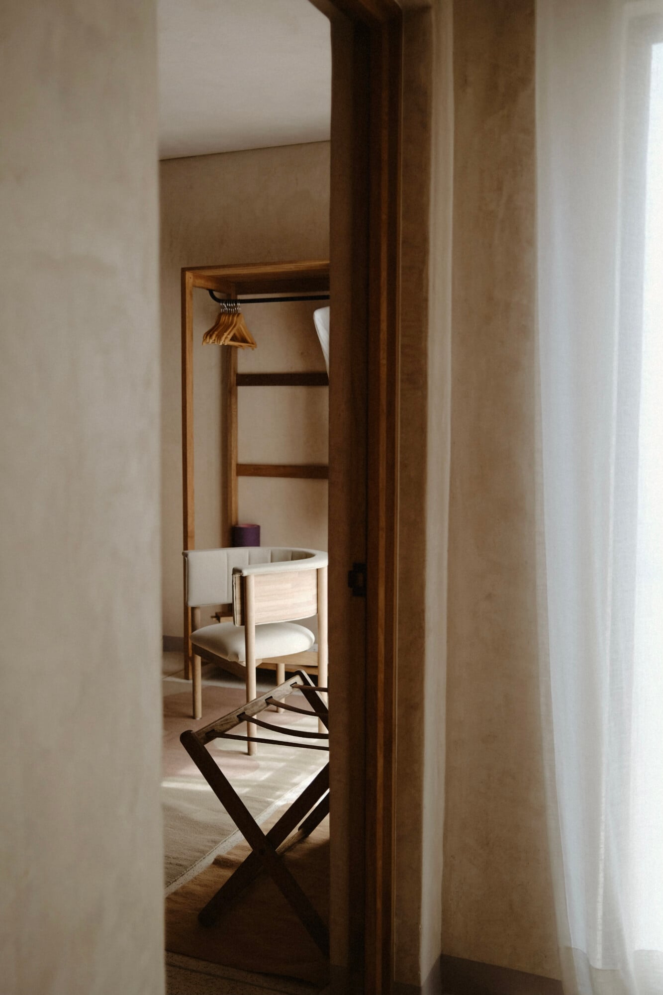 Casa Antonieta, Mexico — Minimalist bedroom closet with wooden frame, white linen chair, brass hangers, and natural light from sheer curtains.