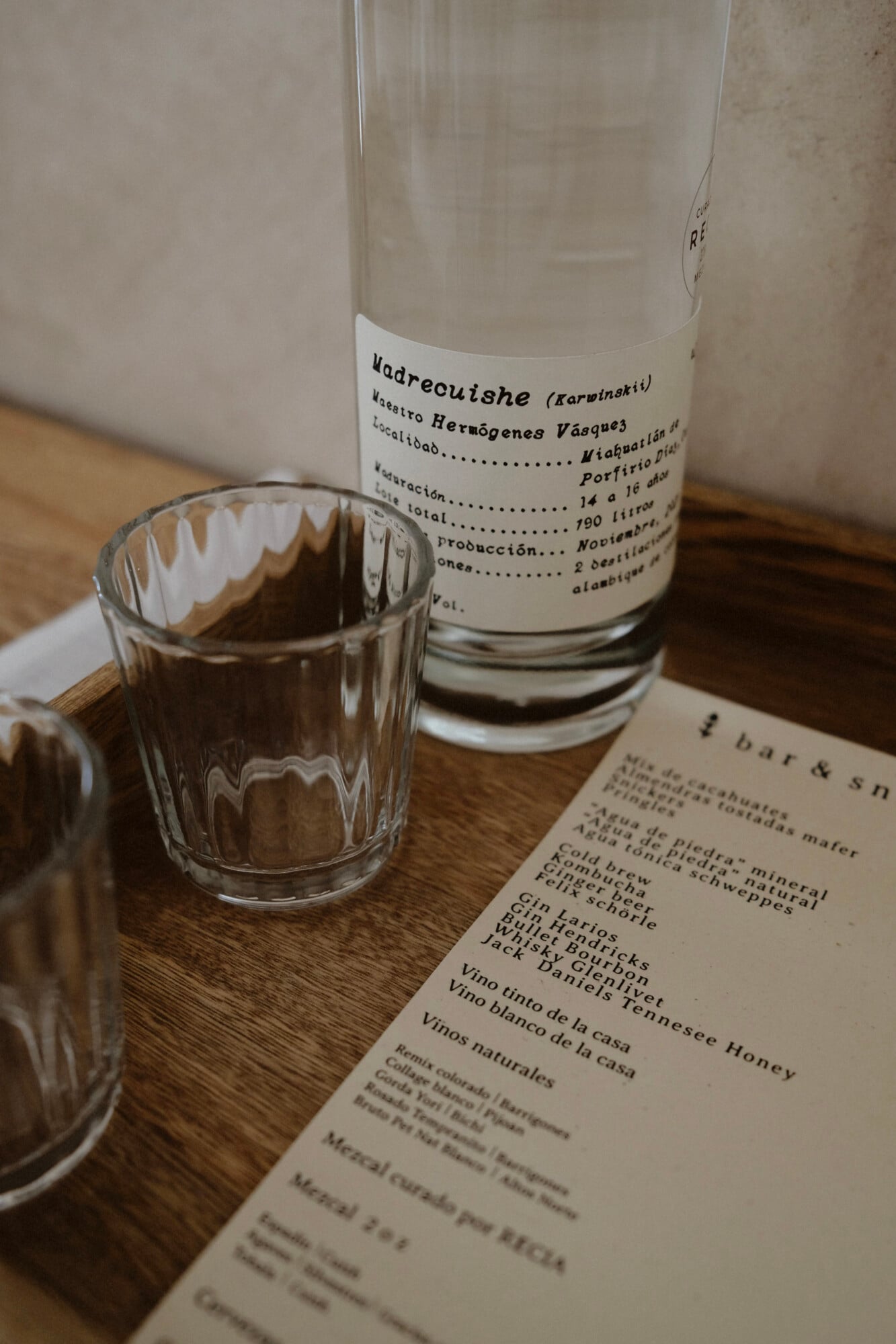 Casa Antonieta, Mexico — Madrecuishe mezcal bottle beside filled glass and bar menu on wooden surface, natural light.