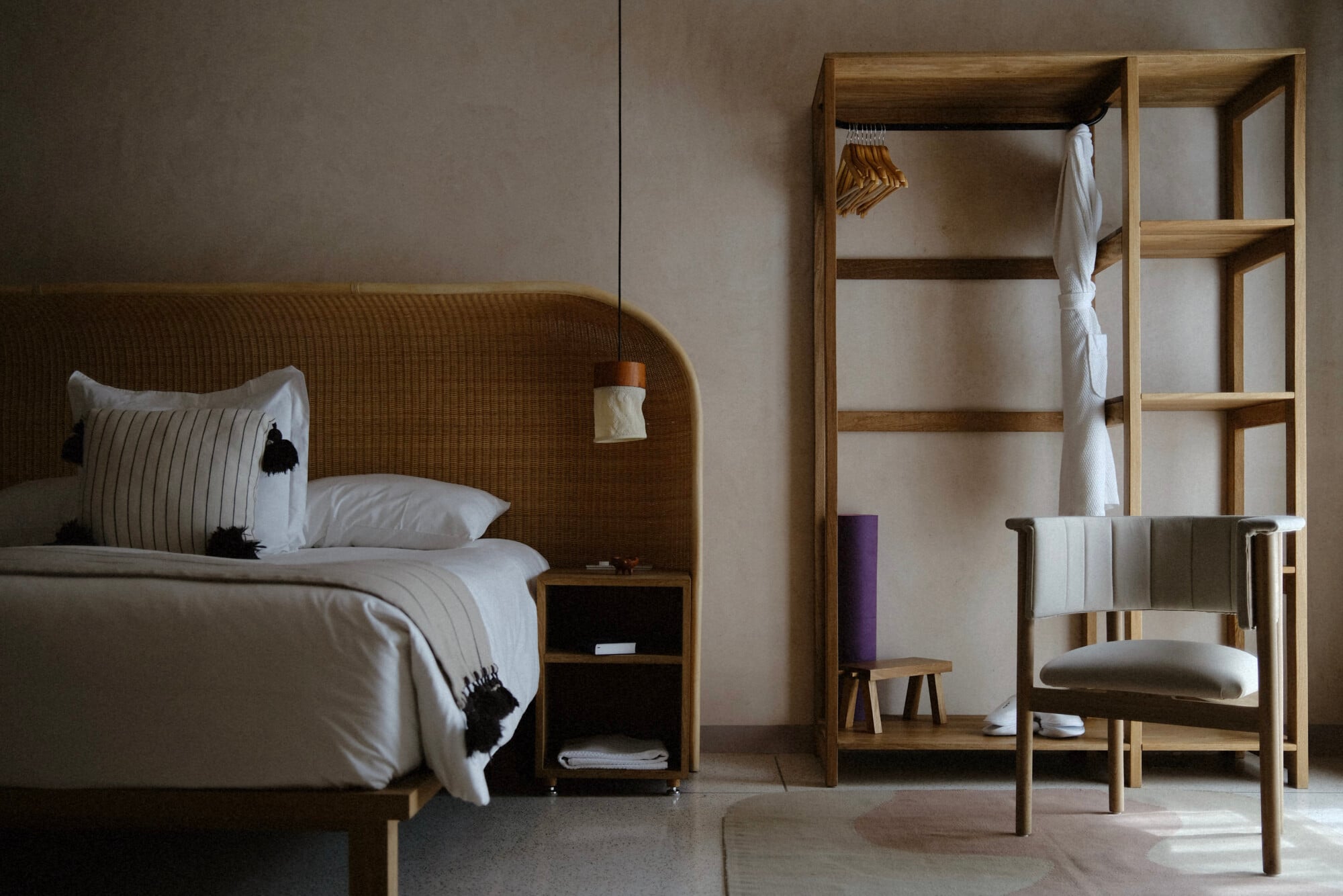 Casa Antonieta, Mexico — Minimalist bedroom with woven rattan headboard, natural wood wardrobe, linen bedding, and wooden chair in warm neutral palette.