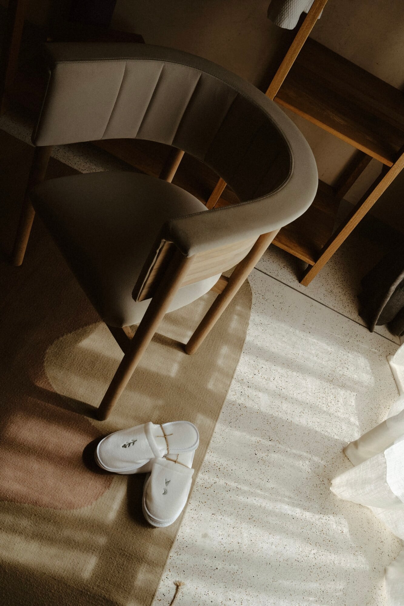 Casa Antonieta, Mexico — Minimalist hotel room with natural light, linen chair, wooden furniture, concrete flooring, and white slippers on woven rug.
