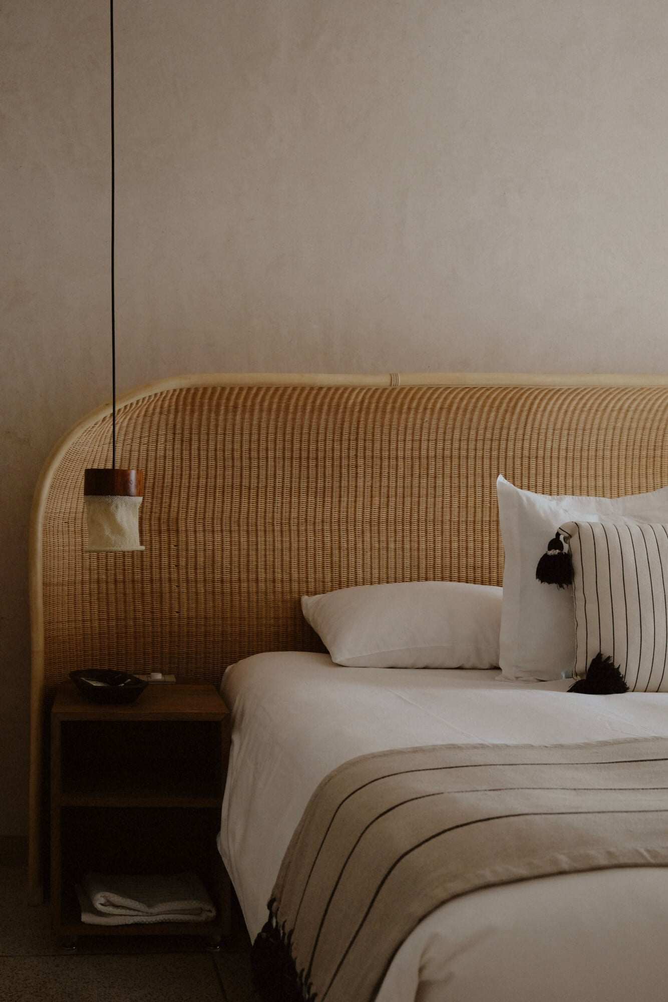 Casa Antonieta, Mexico — Minimalist bedroom with woven rattan headboard, cream linens, striped throw blanket, pendant light, and dark wood nightstand.
