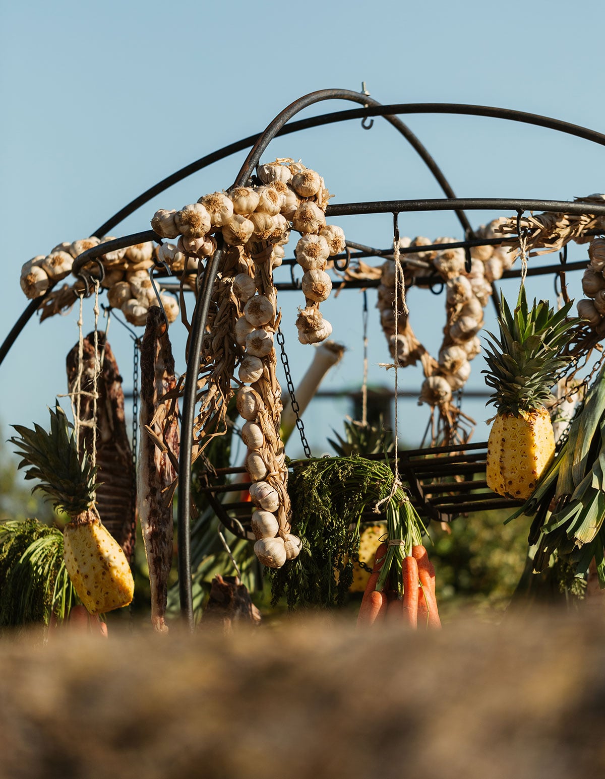 Casa Maca, Balearic Islands — Dried garlic braids and pineapples hanging from wrought-iron garden arbor against clear sky, rustic Mediterranean aesthetic.