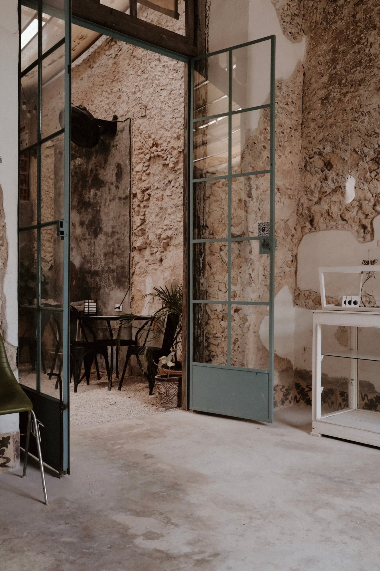 Café Lavé, Mexico — Industrial steel-framed glass doors open to textured stone courtyard with black furniture and natural light filtering through weathered walls.