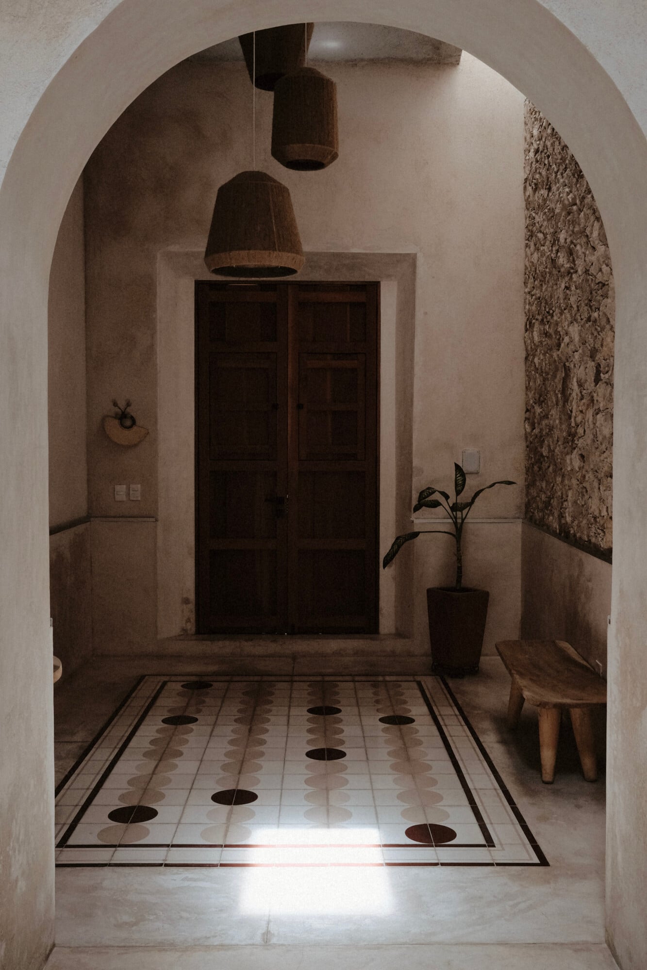 Casa Lohr, Mexico — Arched entryway with geometric tile floor, dark wooden doors, woven pendant lights, and textured stone wall detail in minimalist interior.