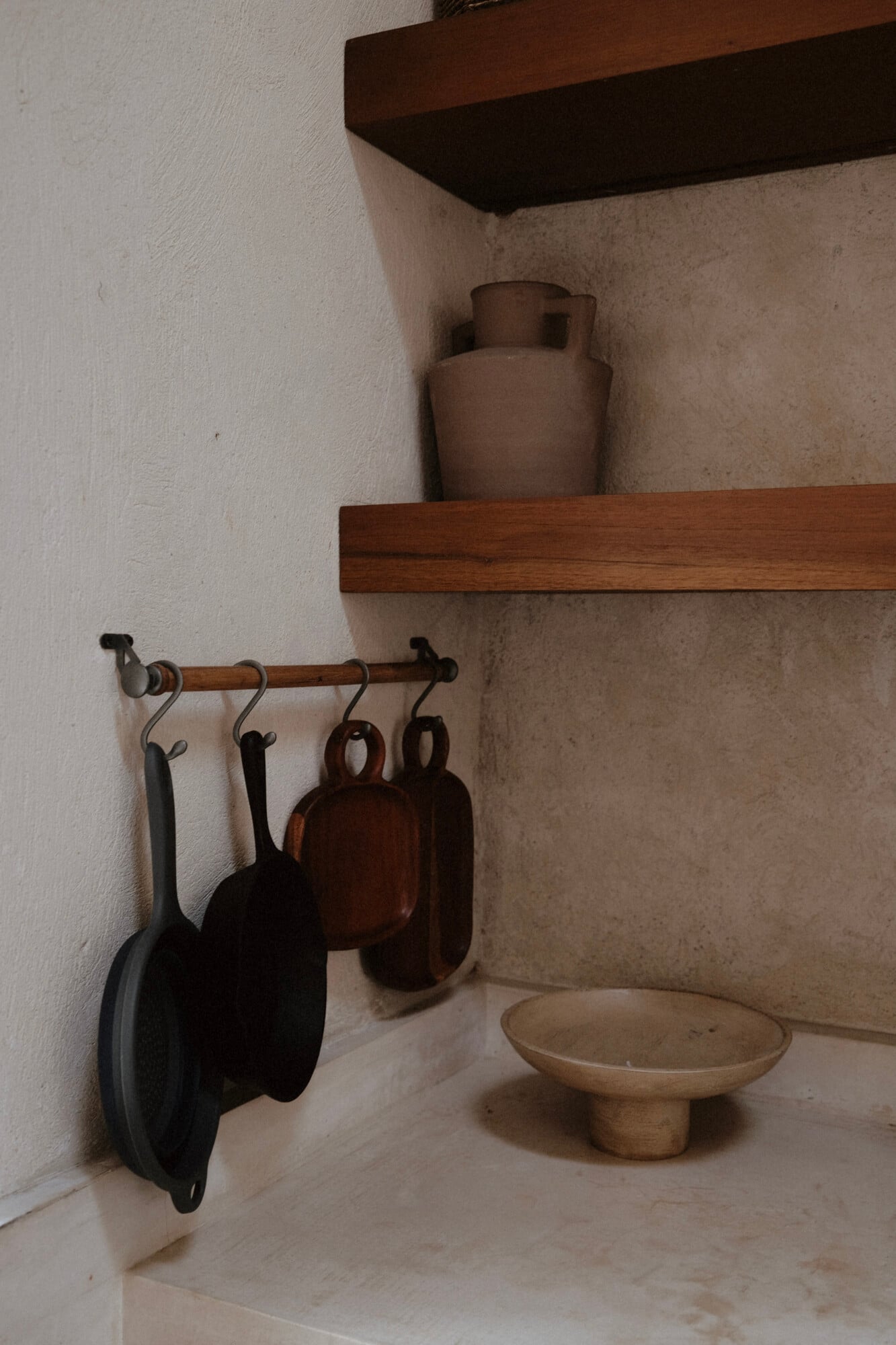 Casa Lohr, Mexico — Rustic kitchen alcove with wooden shelves, clay vessels, cast iron cookware hanging from rod, and terracotta bowl on stone countertop.