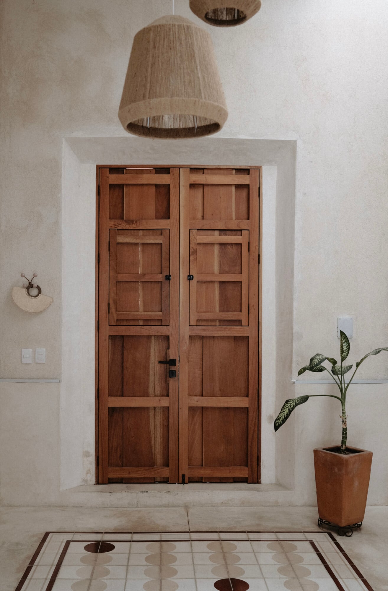 Casa Lohr, Mexico — Wooden double doors with glass panels in whitewashed entry hall, pendant lamp, potted plant, geometric tile floor.