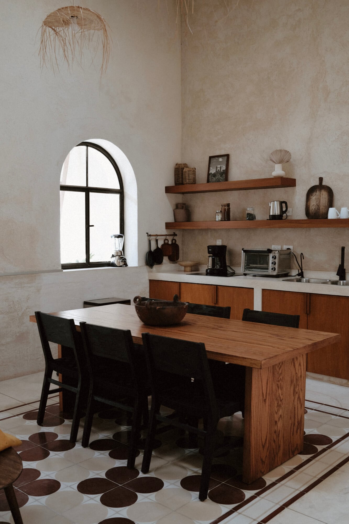 Casa Lohr, Mexico — Minimalist kitchen and dining room with arched window, wooden table, black chairs, floating shelves, terracotta tile floor, and natural light.