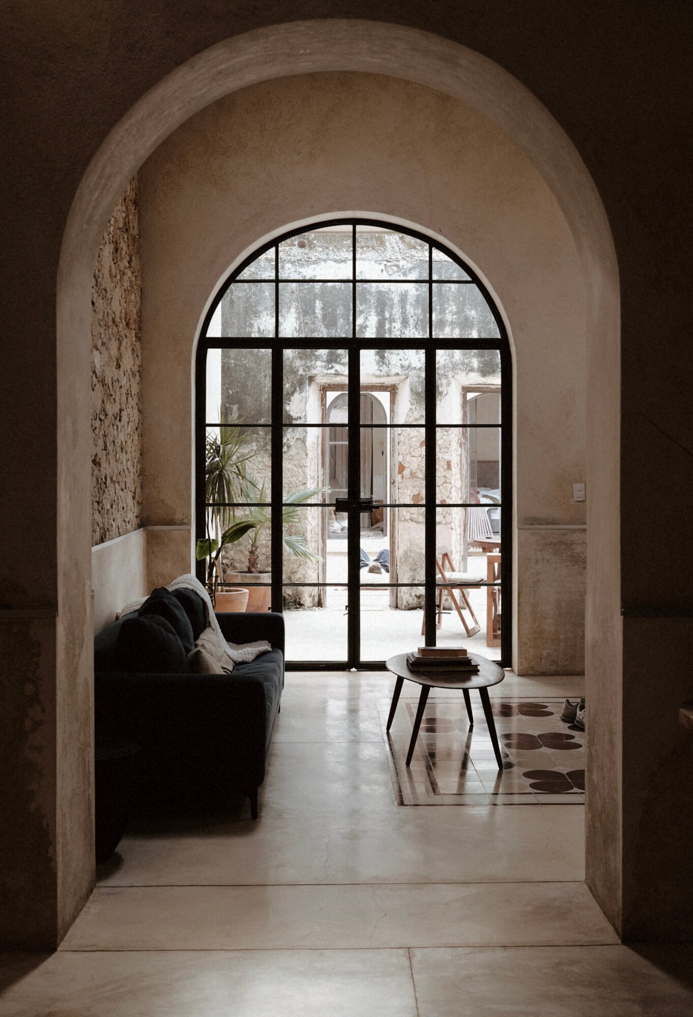 Casa Lohr, Mexico — Arched stone doorway framing black-gridded glass doors opening to sunlit courtyard with minimal furnishings and aged stucco walls.