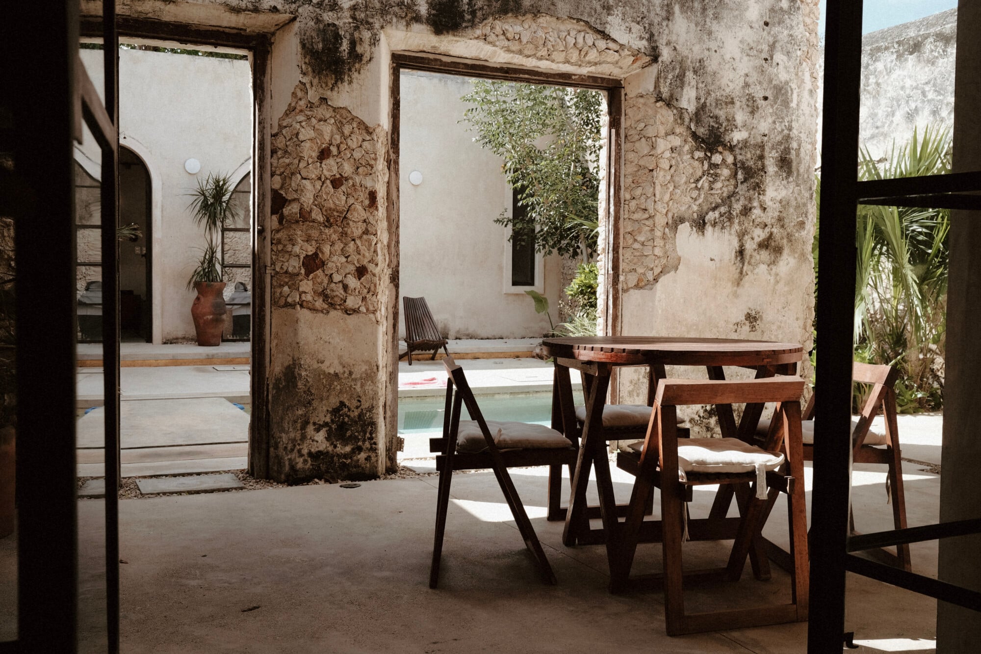 Casa Lohr, Mexico — Rustic concrete courtyard with wooden dining table and chairs, weathered stone columns, pool view, and tropical plants in natural light.
