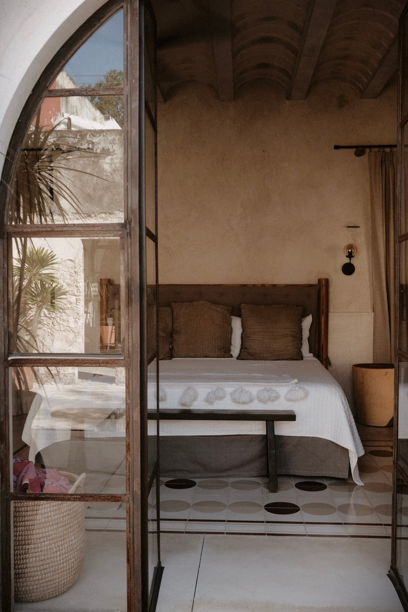 Casa Lohr, Mexico — Minimalist bedroom with arched ceiling, wooden bed frame, white linens, earth-toned pillows, patterned tile floor, palm fronds visible through rustic glass door.