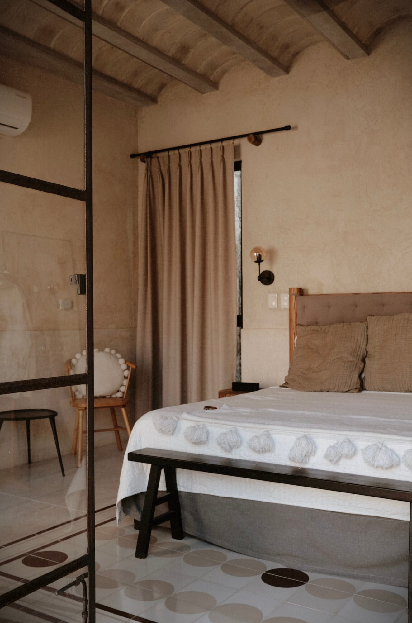 Casa Lohr, Mexico — Minimalist bedroom with black-framed glass partition, natural linen curtains, exposed wooden ceiling beams, and terrazzo flooring with circular inlay detail.