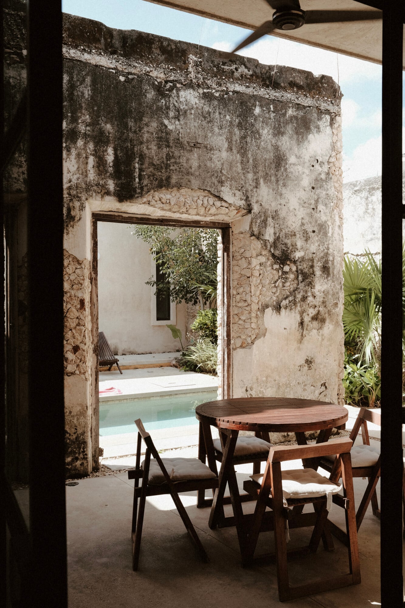 Casa Lohr, Mexico — Weathered stone courtyard with wooden dining table, chairs, pool beyond, tropical plants, and minimal modern furnishings in austere colonial architecture.