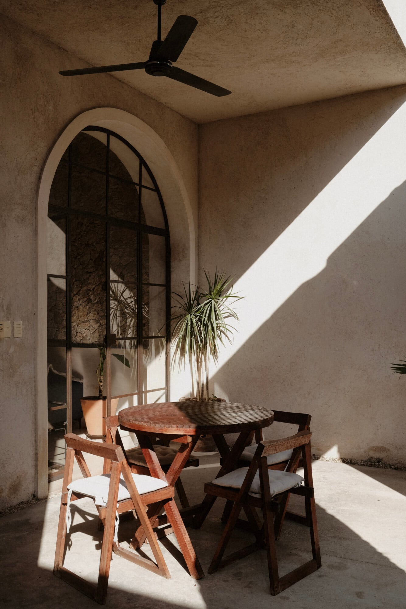 Casa Lohr, Mexico — Arched doorway with black steel frame, wooden dining table, mid-century chairs, potted palm, concrete walls, ceiling fan, natural light casting shadows.