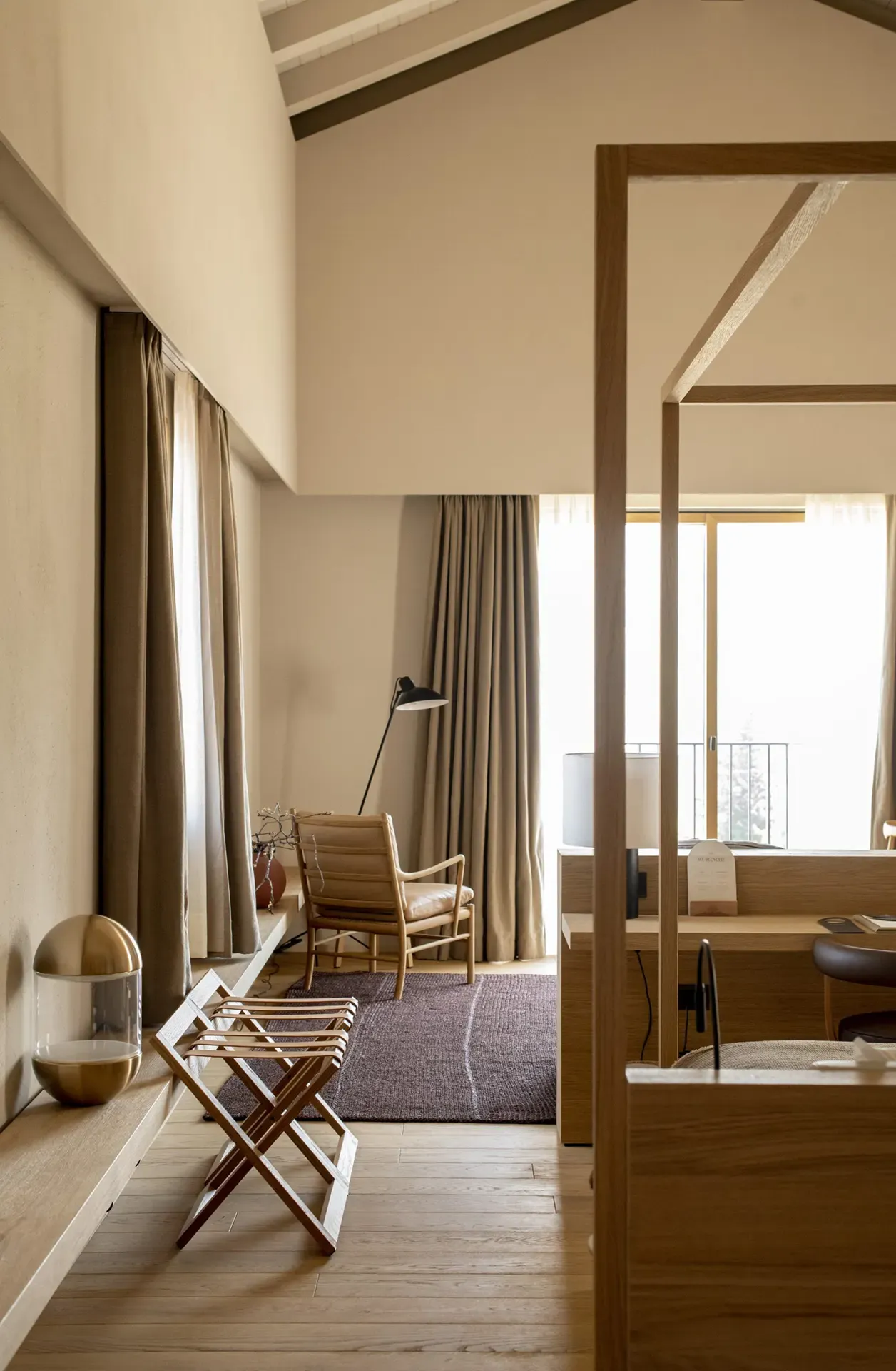 Casa di Langa, Europe — Minimalist hotel bedroom with wooden four-poster bed, mid-century lounge chair, floor-to-ceiling windows, neutral palette, natural light, and modern architectural details.
