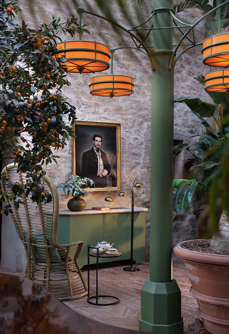 A courtyard at Castello di Reschio glows under orange pendant lamps, framed by weathered stone walls, potted plants, and a portrait in gilt.