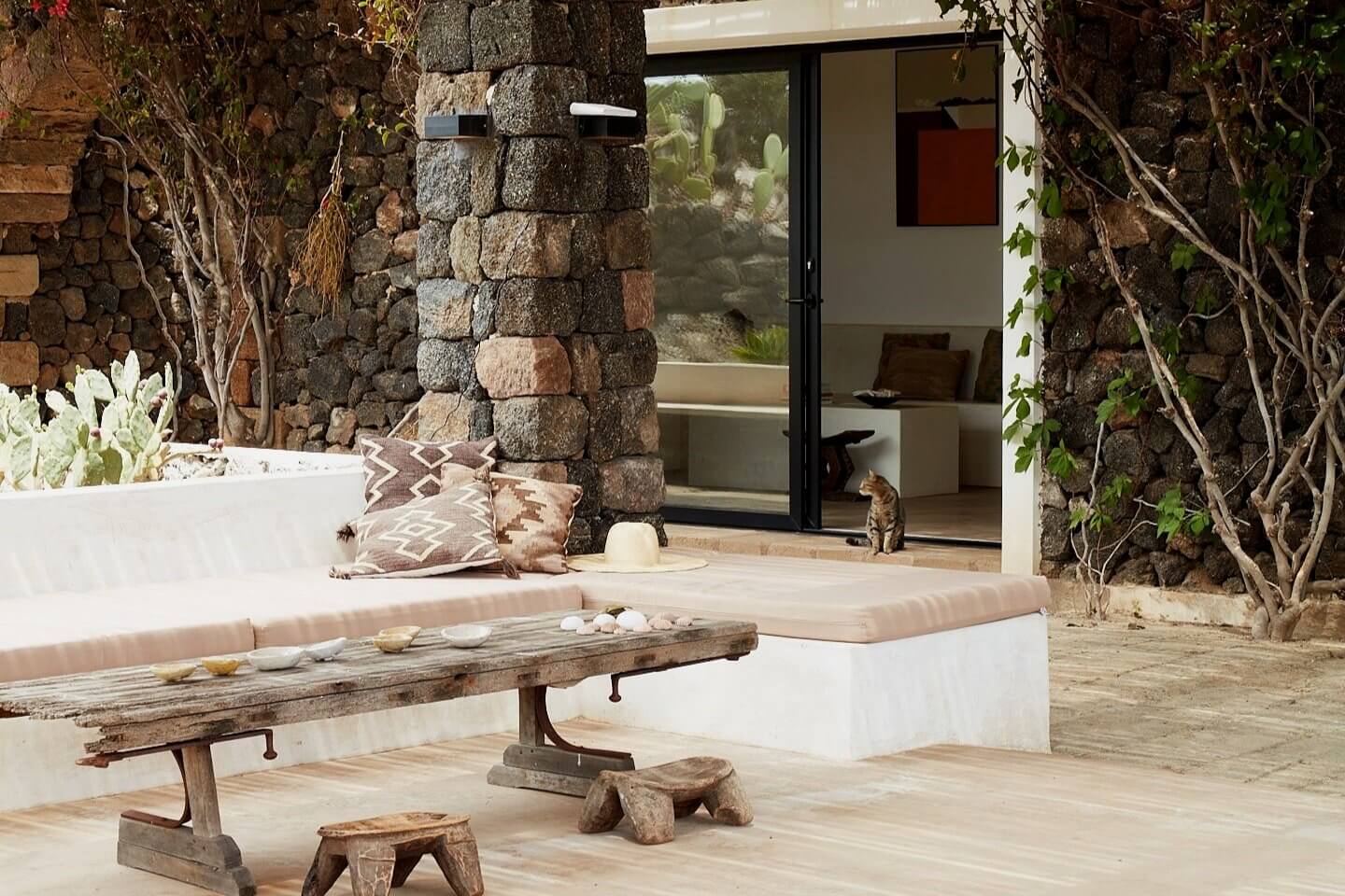 Parco Dei Sesi, Europe — Courtyard with stone columns, whitewashed seating, weathered wooden bench, trailing vines, and modern glass doors opening to interior spaces.