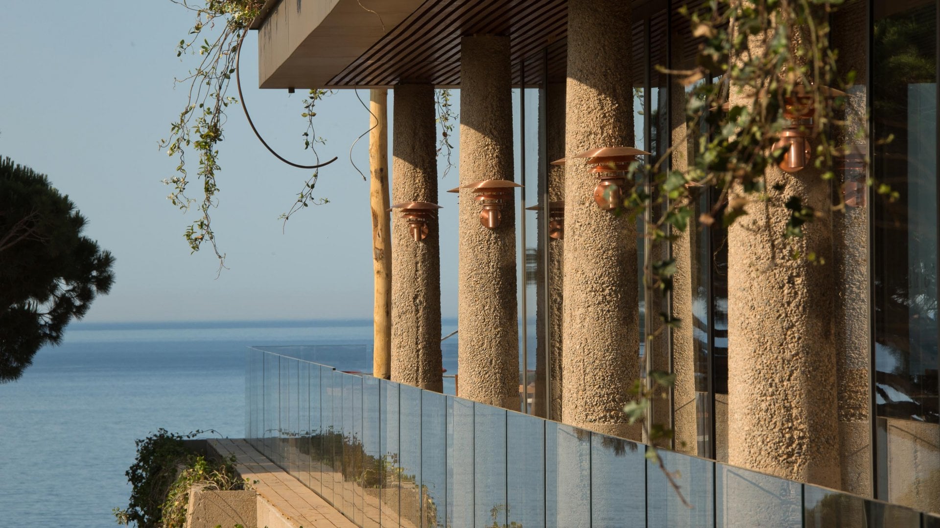 Lily of the Valley, Europe — Coastal modernist terrace with textured stone columns, glass balustrade, hanging ivy, and tranquil sea views under clear sky.