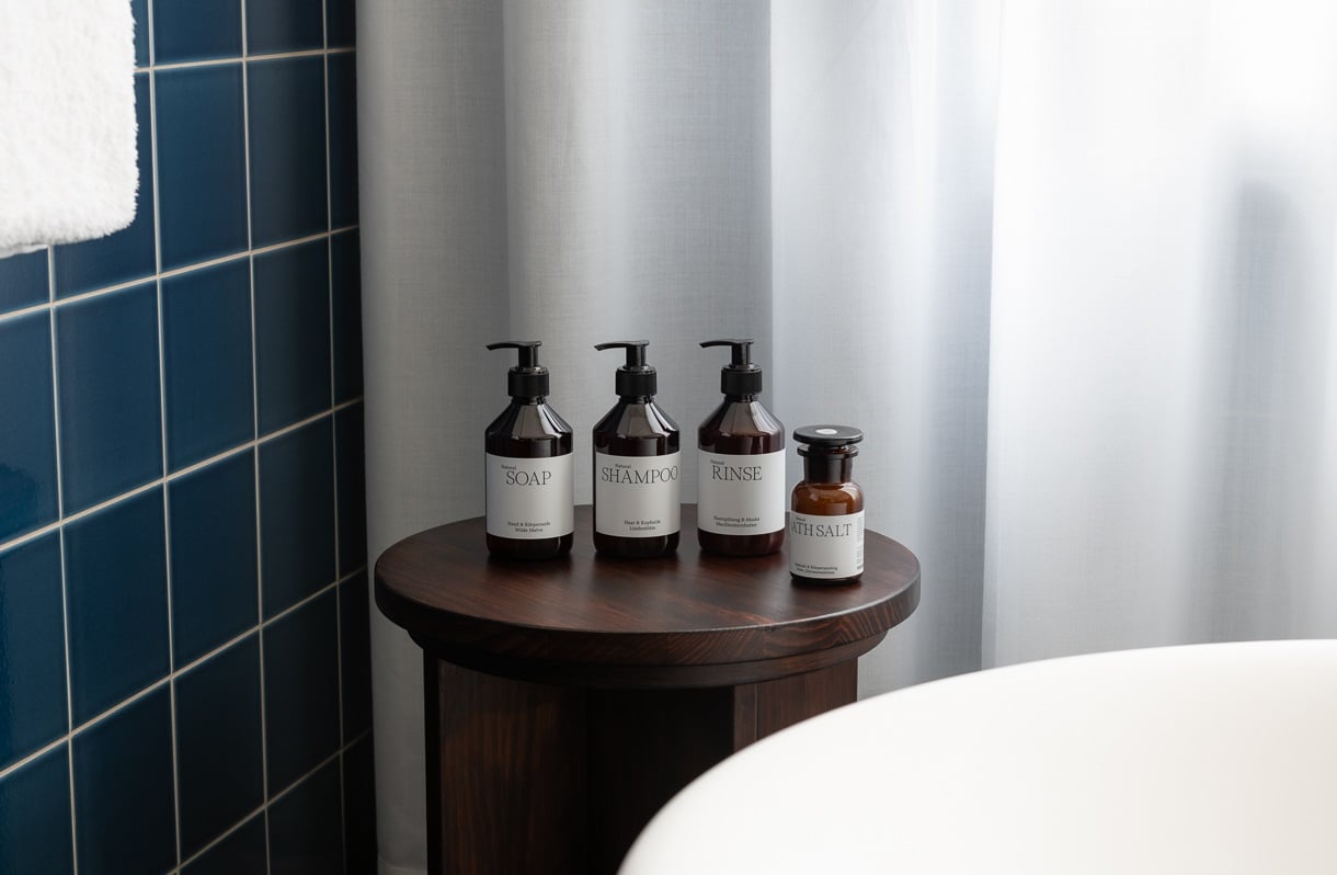 Château Royal, Europe — Dark-tiled bathroom corner with dark wood pedestal table displaying luxury toiletries in amber glass bottles with pump dispensers, minimalist aesthetic.