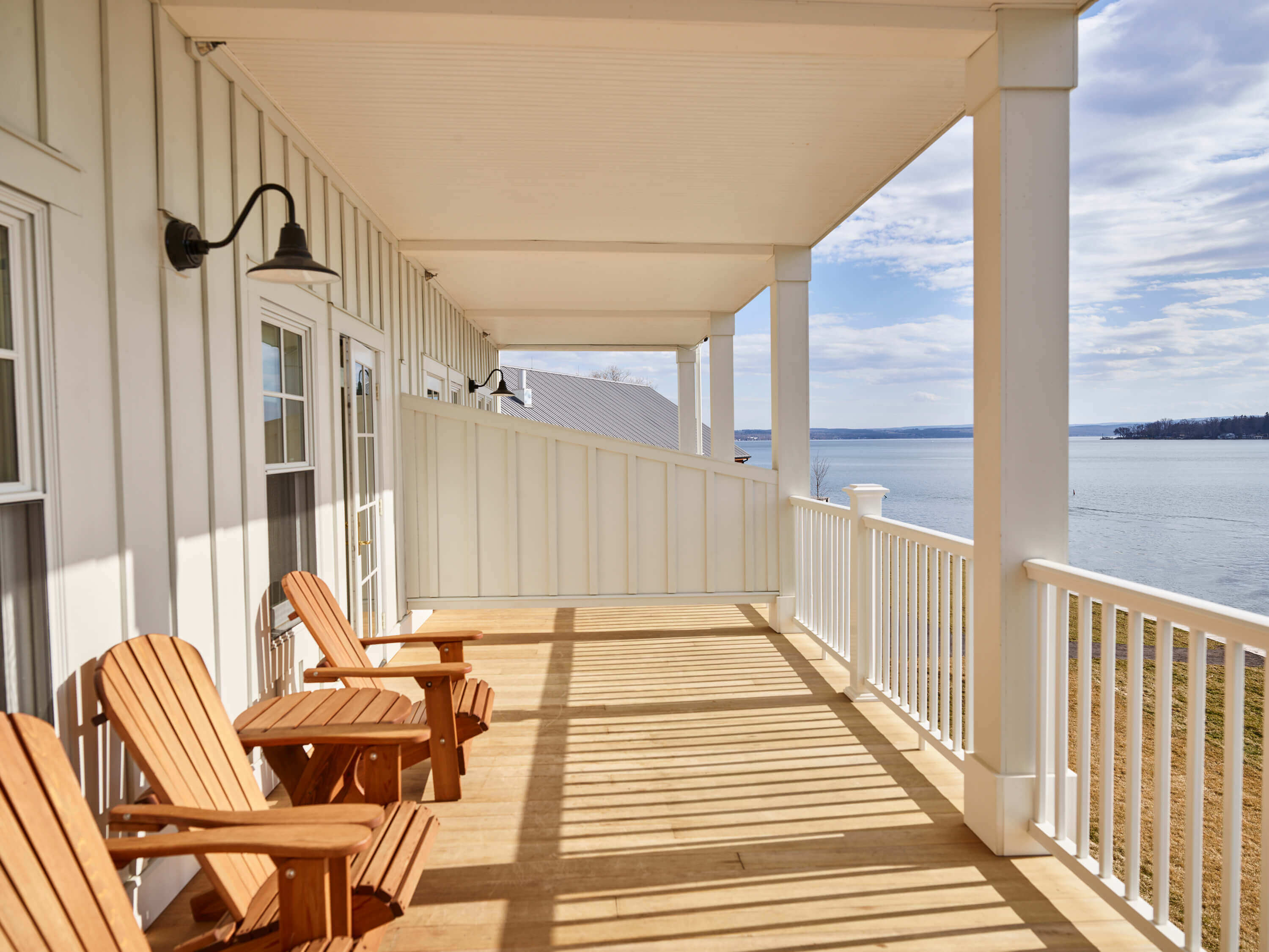 The Lake House, North America — Covered waterfront deck with cedar Adirondack chairs, white railings, and black pendant lights overlooking calm bay waters.