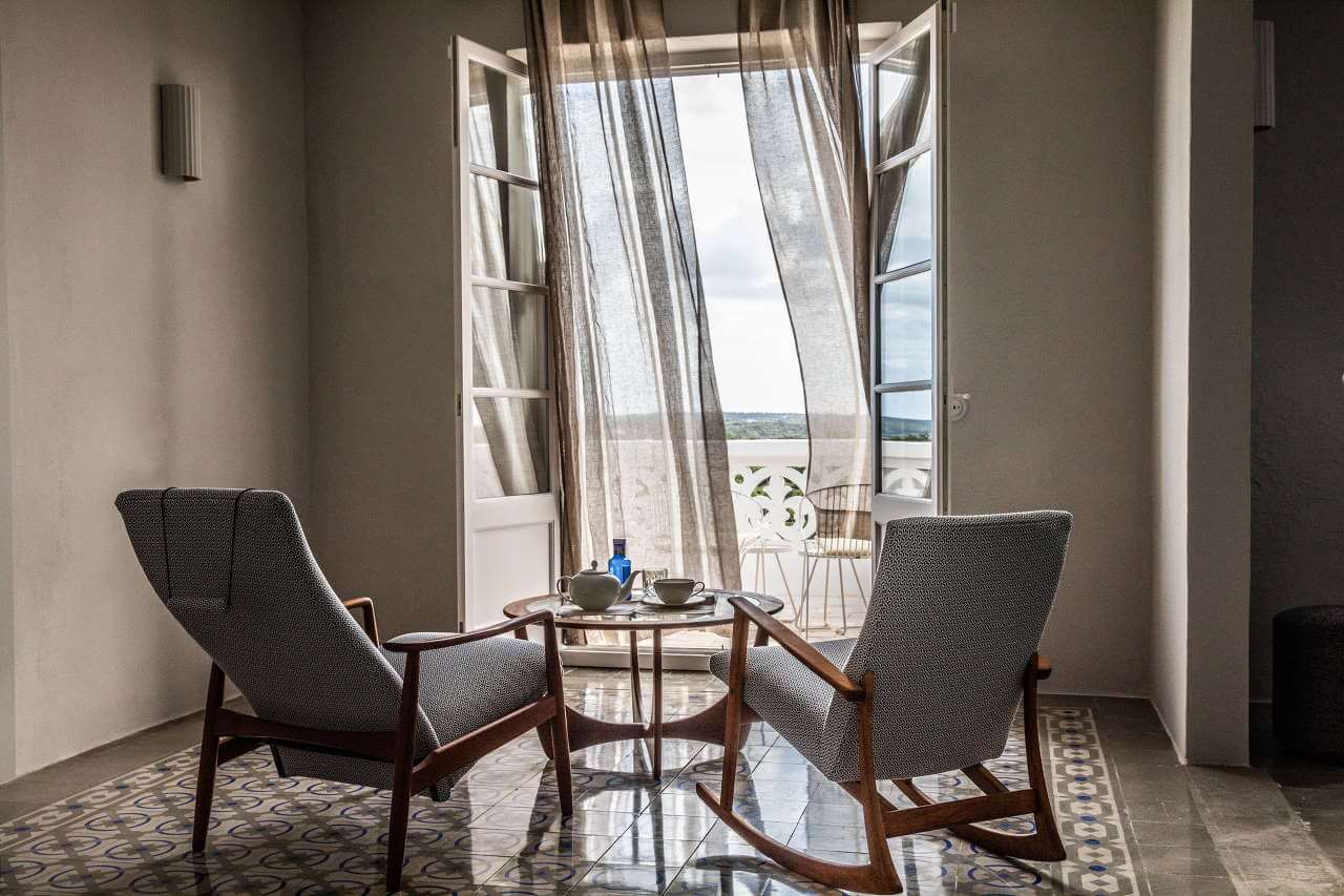 Es Bec d’Aguila — Es Bec d'Aguila, Balearic Islands, Spain — Mid-century lounge chairs facing open French doors with linen curtains, coastal views, polished stone floors, natural light.