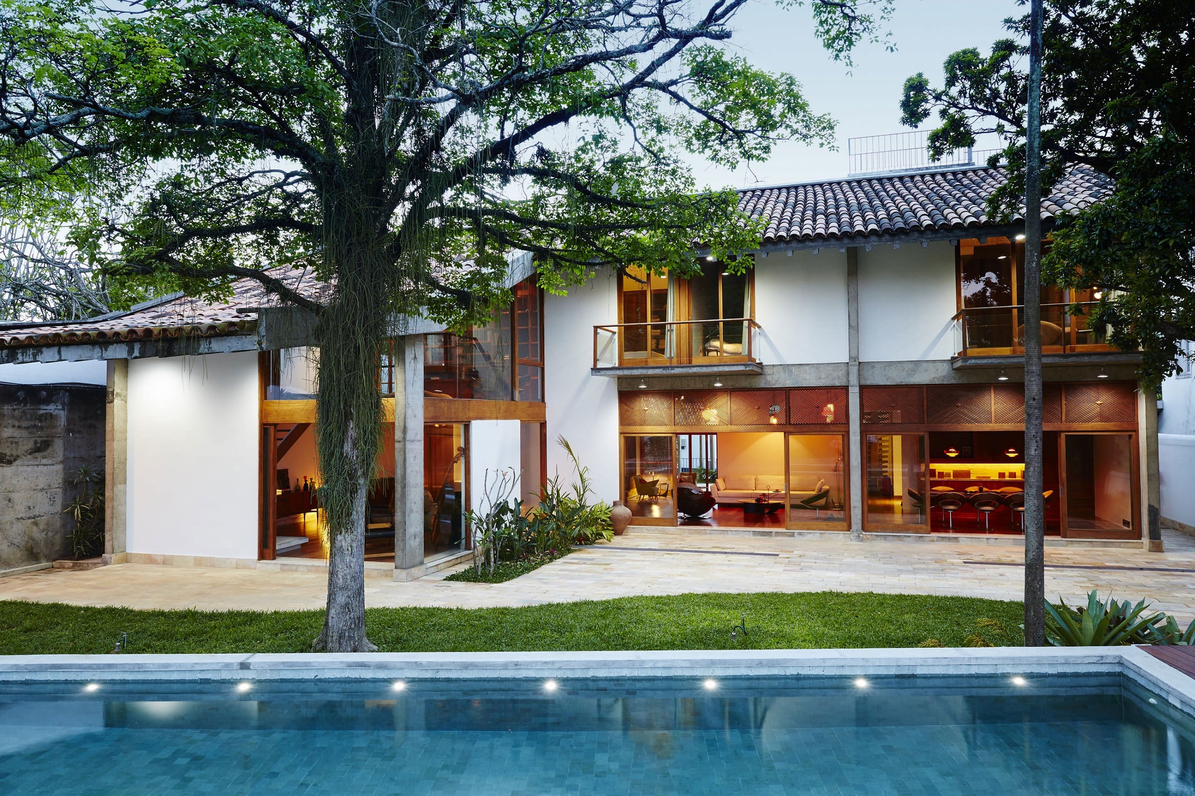 Chez Georges, South America — modernist courtyard with illuminated pool, whitewashed walls, wooden shutters, mature trees, warm interior lighting at dusk.