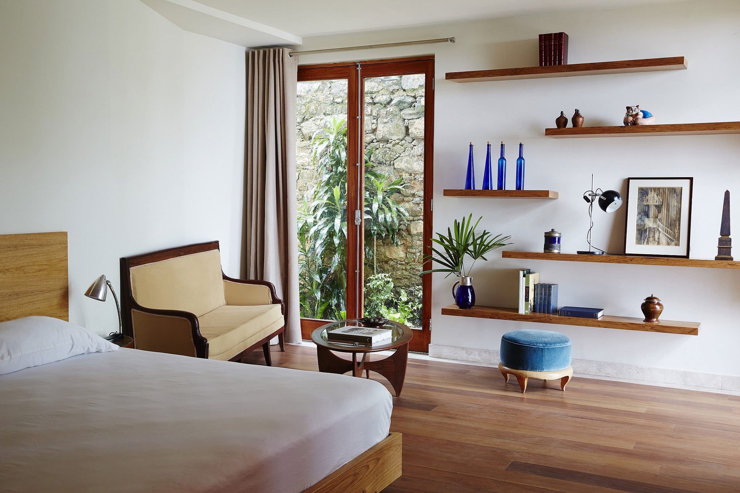 Chez Georges, South America — Modernist bedroom with walnut floating shelves, cream armchair, blue velvet ottoman, wooden doors opening to stone garden, warm ambient lighting.