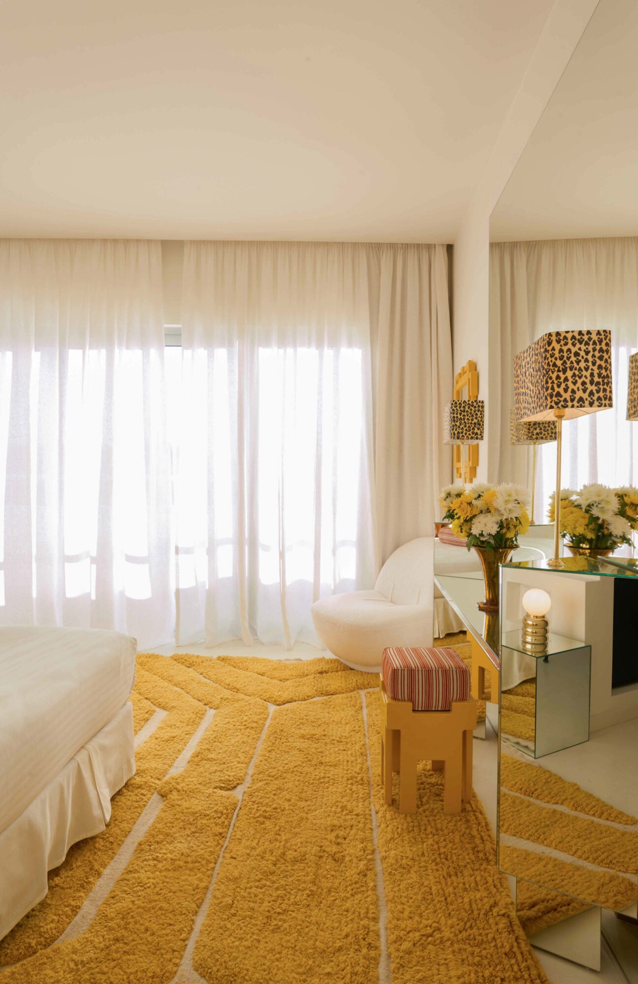 Apollo Palm, Europe — Sunlit hotel room with mustard geometric rug, cream furnishings, mirrored vanity, and sheer curtains filtering natural light.