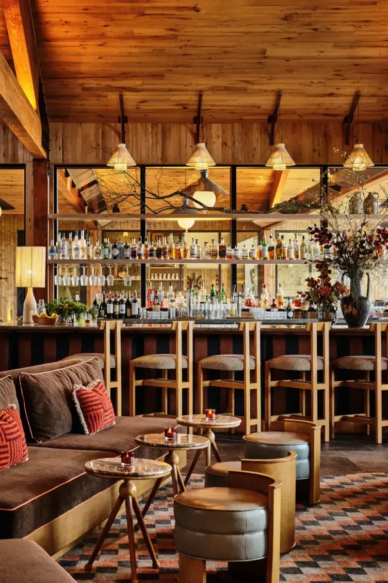 Wildflower Farms, North America — Warm wooden lodge bar with brass pendant lights, extensive spirit collection, leather seating, and mid-century modern furnishings overlooking forest views.