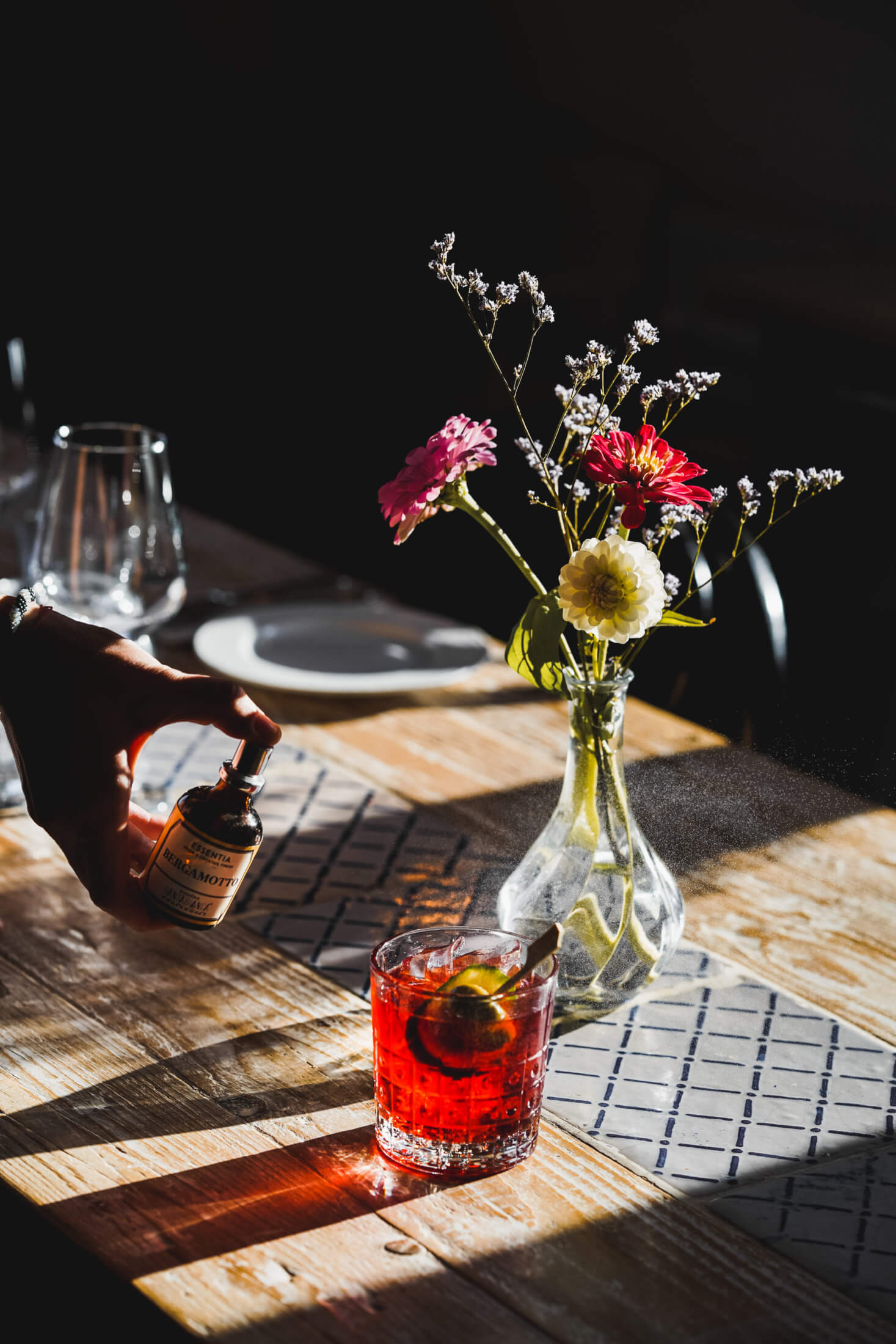 Villa Lena, Europe — Sunlit wooden table set with fresh floral arrangement, cocktail, and place settings against dark interior backdrop.
