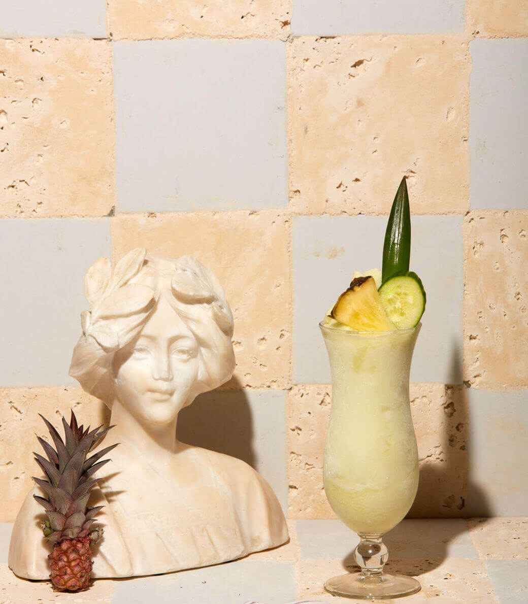 Coconut Club, North America — Pale travertine tile backdrop with classical bust, tropical cocktail garnished with pineapple and cucumber, minimalist luxury bar styling.