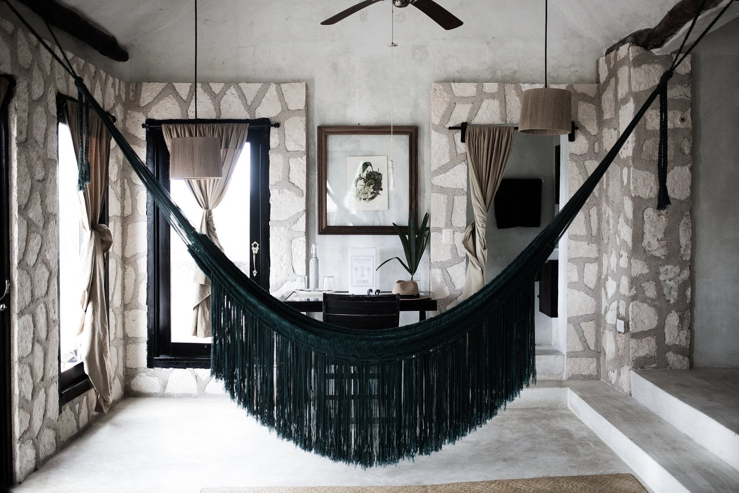 Coqui Coqui Papholchac, Mexico — Dark fringed hammock suspended in stone-walled room with natural light, wooden beams, and botanical artwork.
