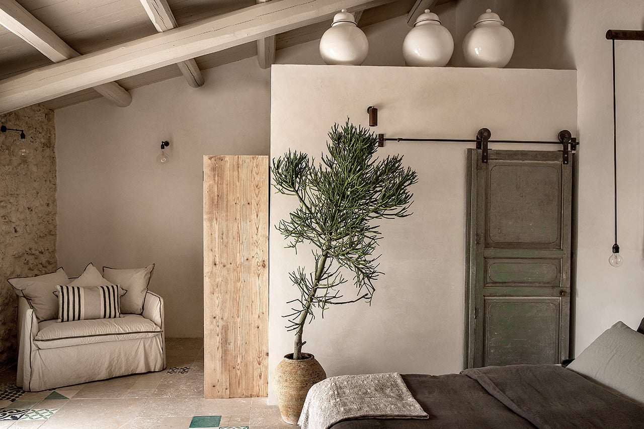 Dimora delle Balze, Europe — Minimalist bedroom with exposed beam ceiling, linen upholstered chair, potted plant, wooden room divider, and vintage barn door sliding hardware on whitewashed walls.
