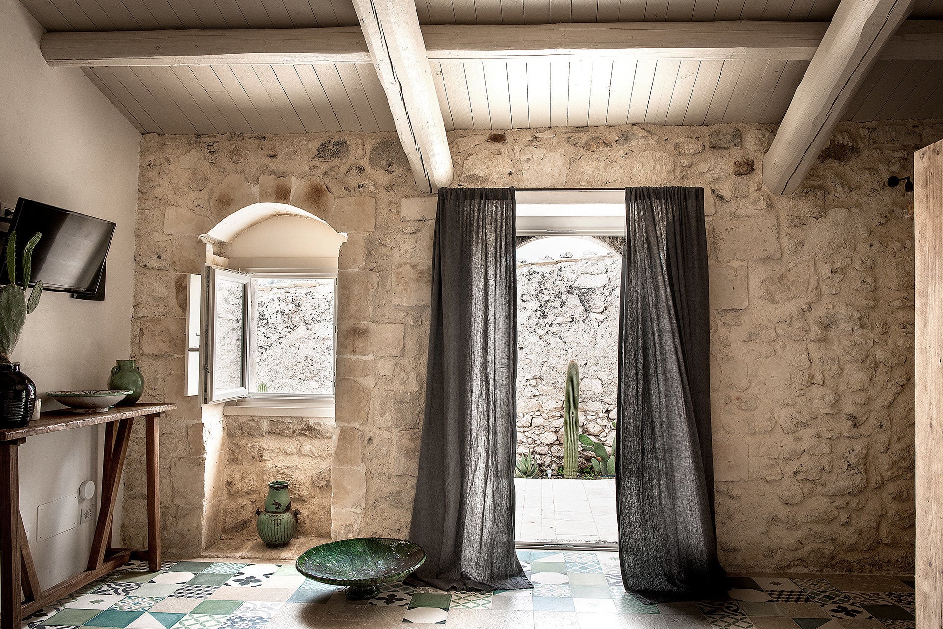 Dimora delle Balze, Europe — Stone-walled room with arched window, dark linen curtains, wooden ceiling beams, ceramic vessels, and geometric tile floor in minimalist luxury design.