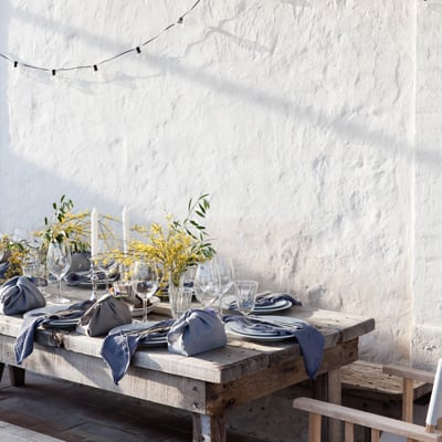 São Lourenço do Barrocal, Portugal — Whitewashed terrace with timber dining table, navy linens, olive branches, string lights, and textured stone wall casting shadows.
