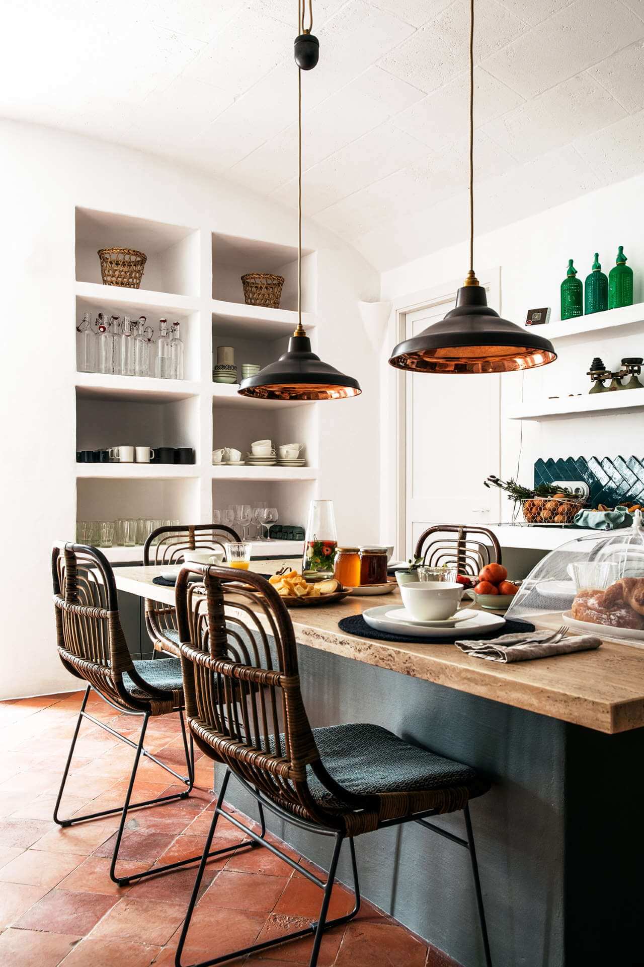 Es Bec d’Aguila — Es Bec d'Aguila, Balearic Islands, Spain — Modern kitchen island with woven rattan chairs, black pendant lights with copper interiors, white built-in shelving, and terracotta tile flooring.