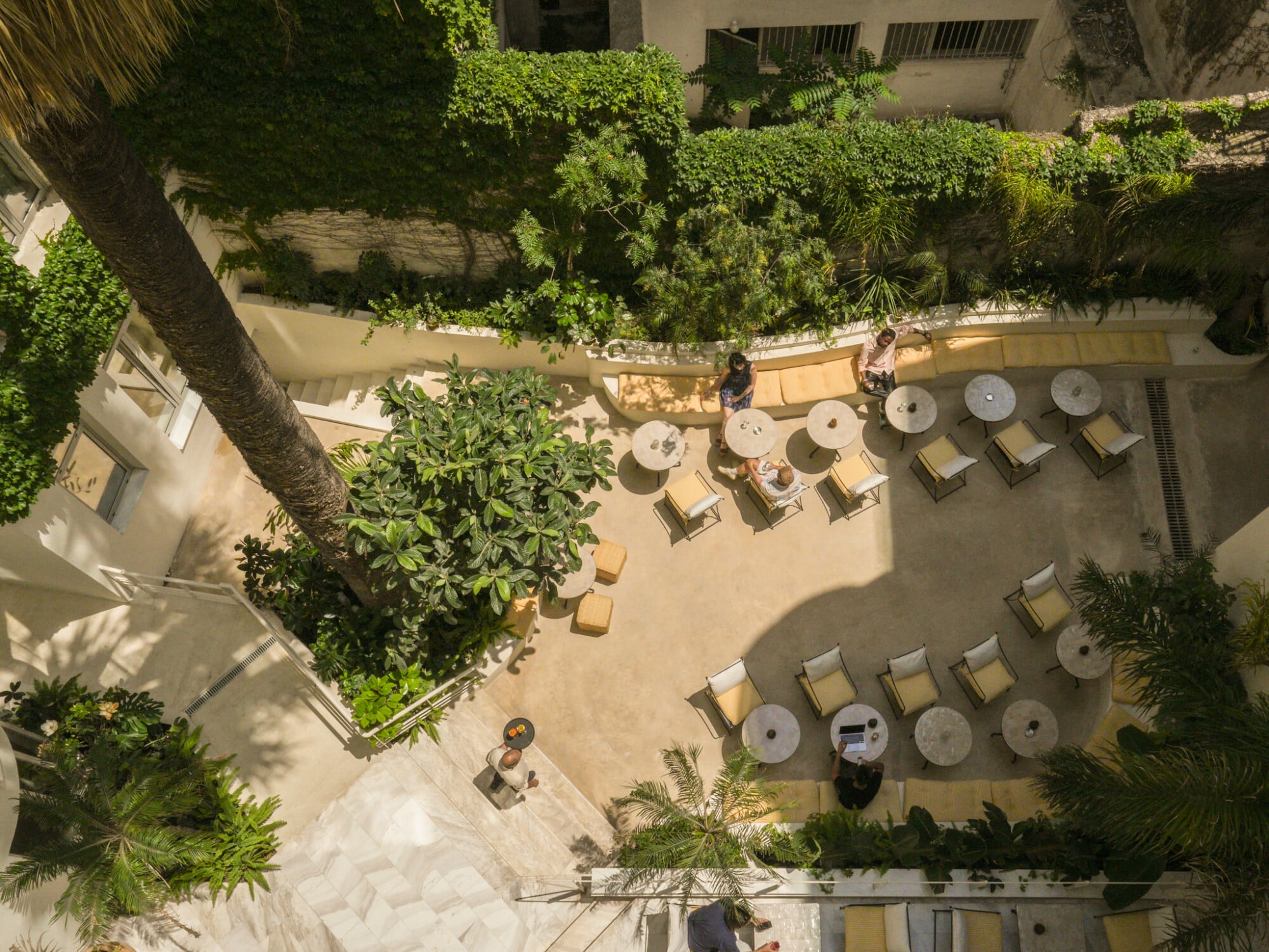 Apollo Palm, Europe — Aerial view of sunlit courtyard with lounge chairs, umbrellas, lush greenery, and Mediterranean architecture.