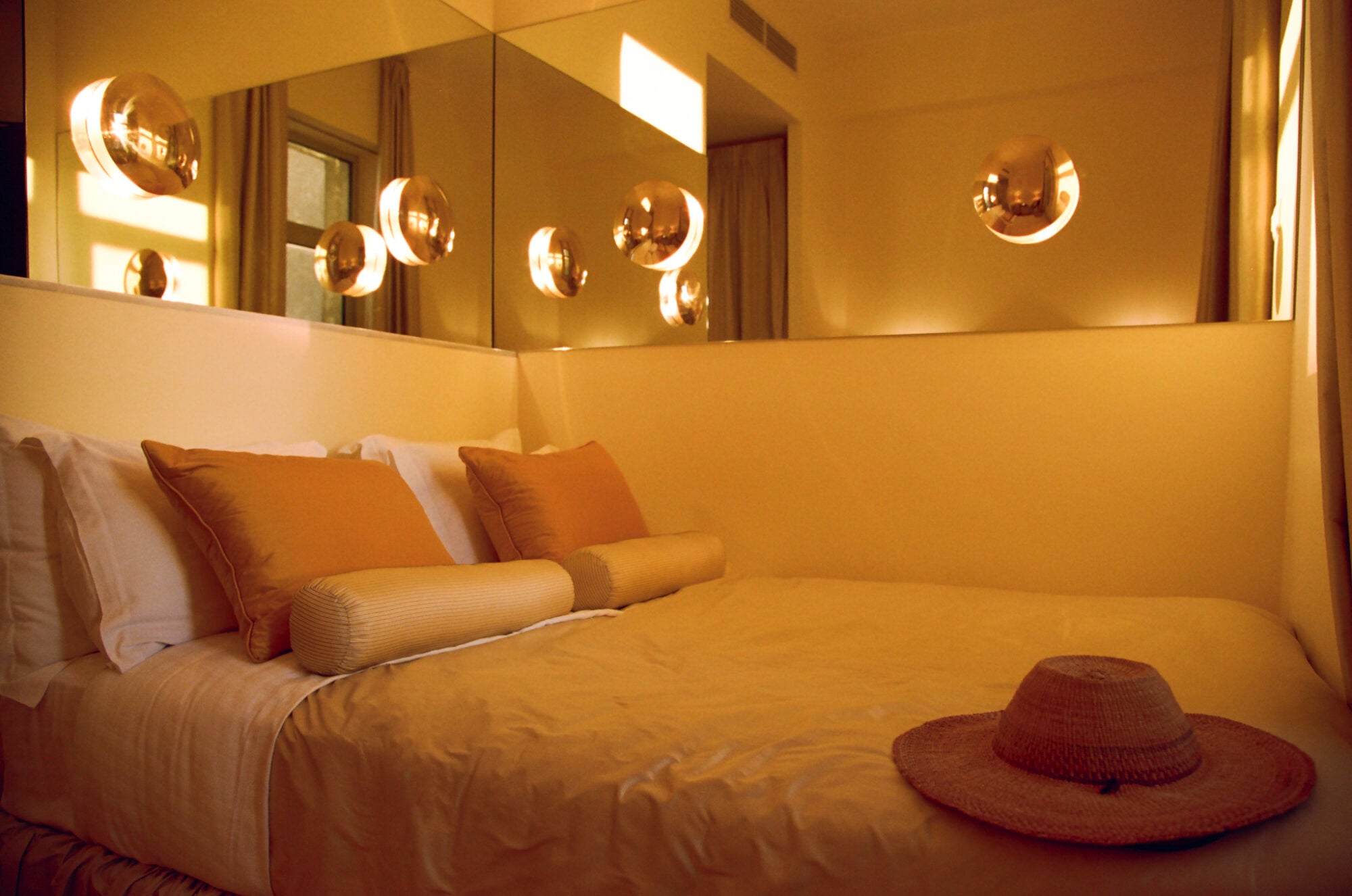 Apollo Palm, Europe — Minimalist hotel bedroom with warm golden walls, mirrored headboard, brass sconces, layered linens, and straw hat on terrazzo floor.