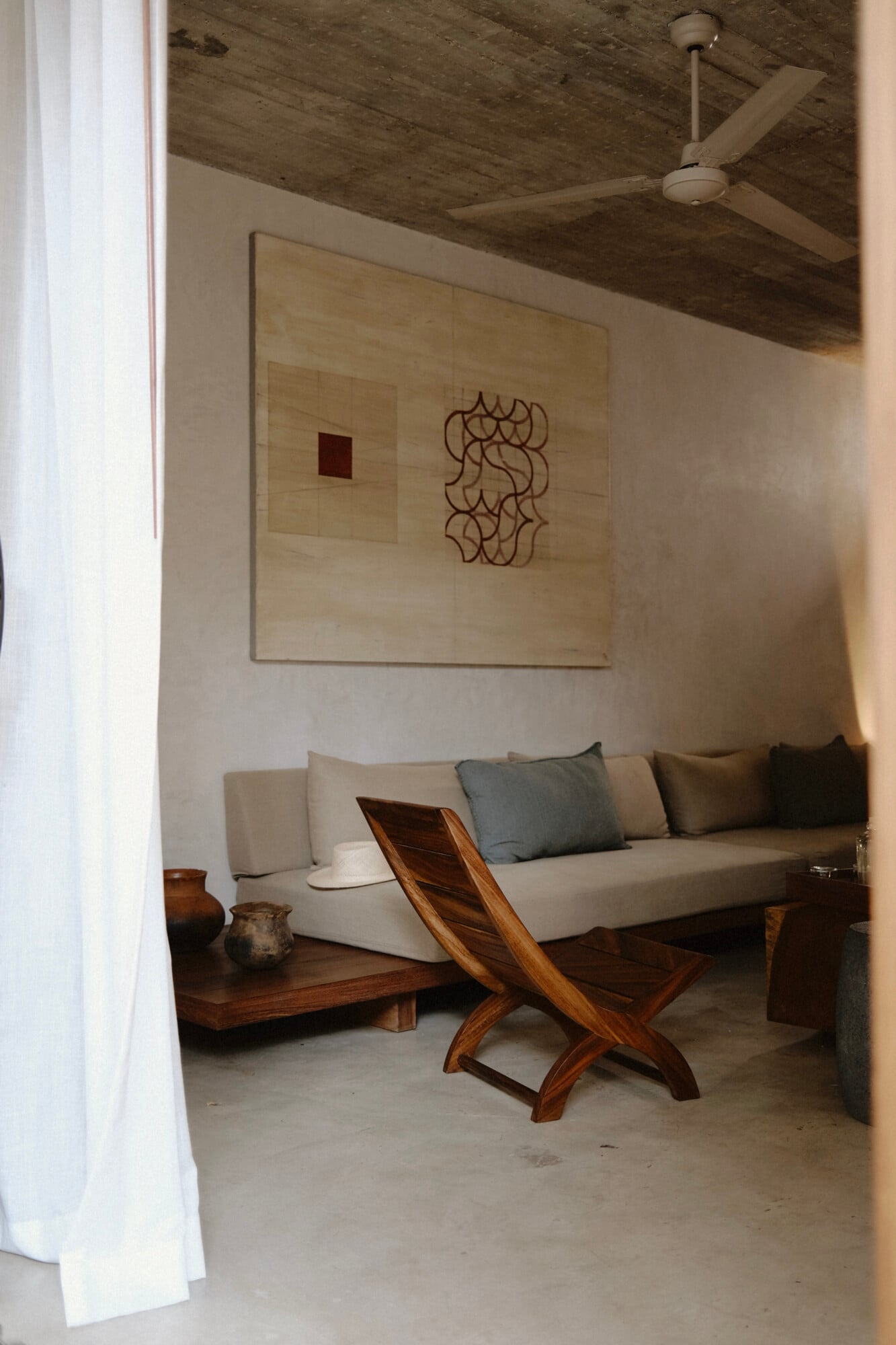 Casa Criollo, Mexico — Minimalist lounge with concrete floor, wooden ceiling, cream sofa, carved wooden chair, and abstract wall art.