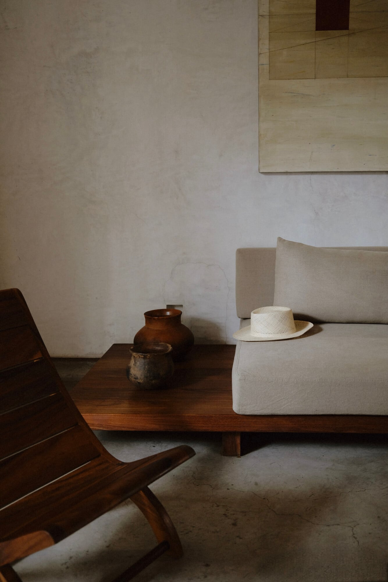 Casa Criollo, Mexico — Minimalist living room with cream linen sofa, dark wooden lounge chair, ceramic vessels, and straw hat on concrete floor