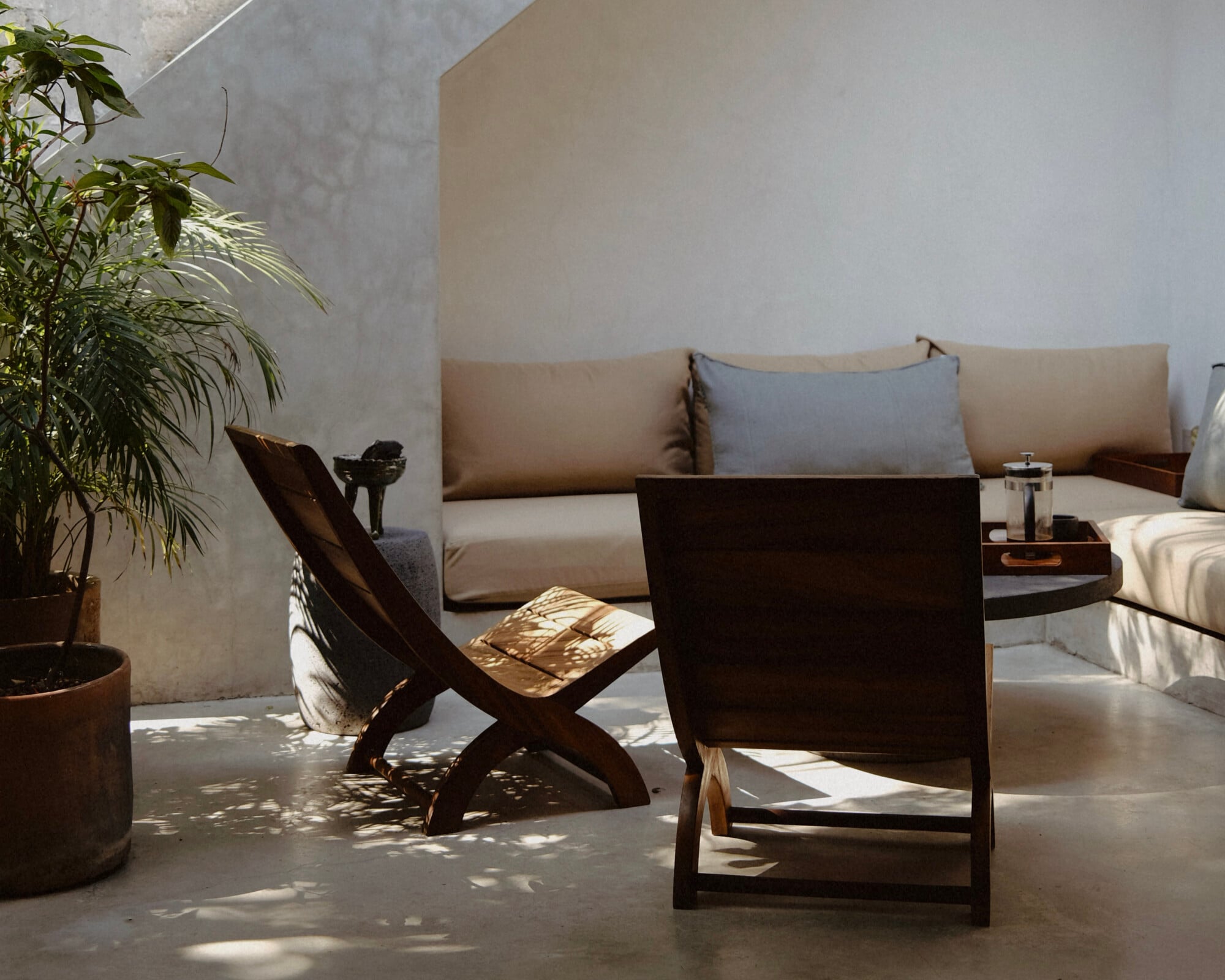 Casa Criollo, Mexico — Minimalist courtyard with concrete walls, dark wood lounge chairs, neutral linen sofa, potted plants, and dappled sunlight casting shadows across polished floor.