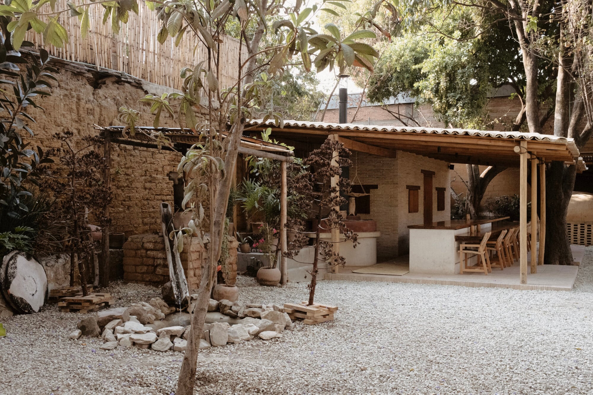 Casa Criollo, Mexico — Courtyard with terracotta brick buildings, wooden pergola, gravel grounds, mature trees, and stone-edged planters creating warm, minimalist hospitality atmosphere.