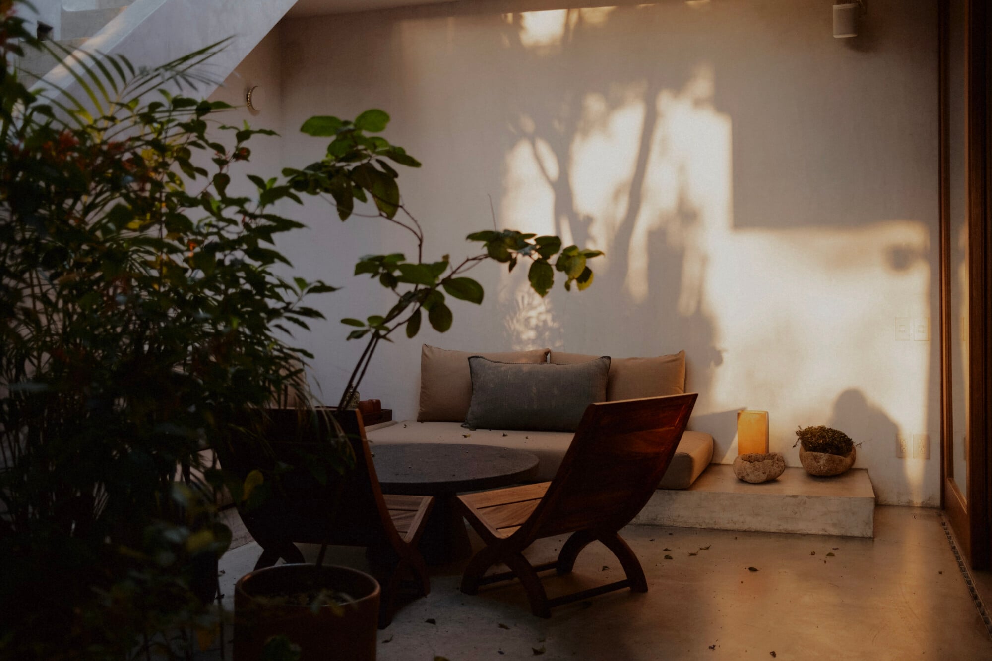 Casa Criollo, Mexico — Sunlit courtyard with wooden lounge chairs, grey cushioned daybed, potted plants casting shadows on whitewashed walls, concrete flooring.