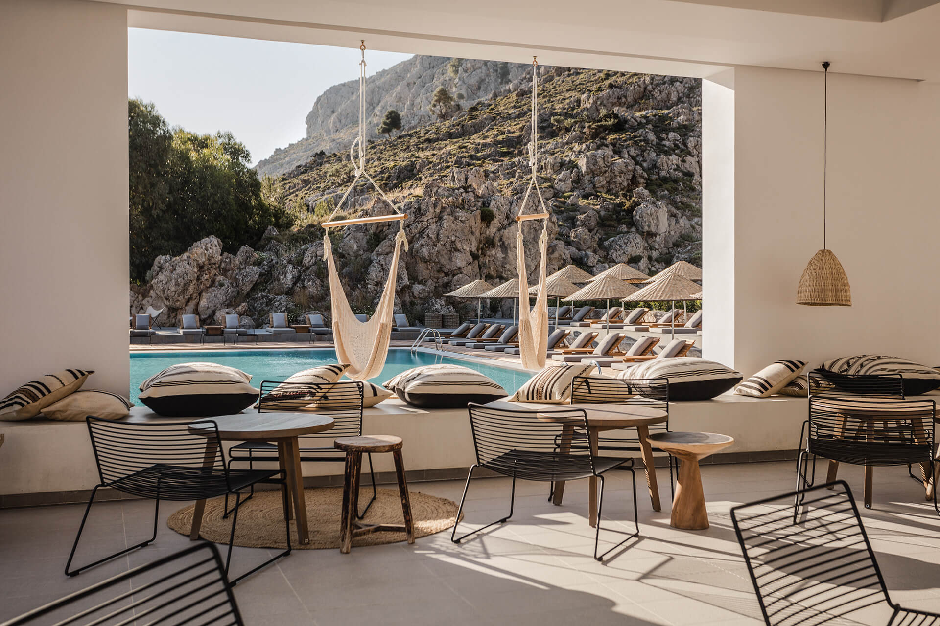 Casa Cook Rhodes, Greece — Minimalist poolside lounge with cream rope swings, striped chairs, natural textiles, and dramatic rocky cliffside backdrop.