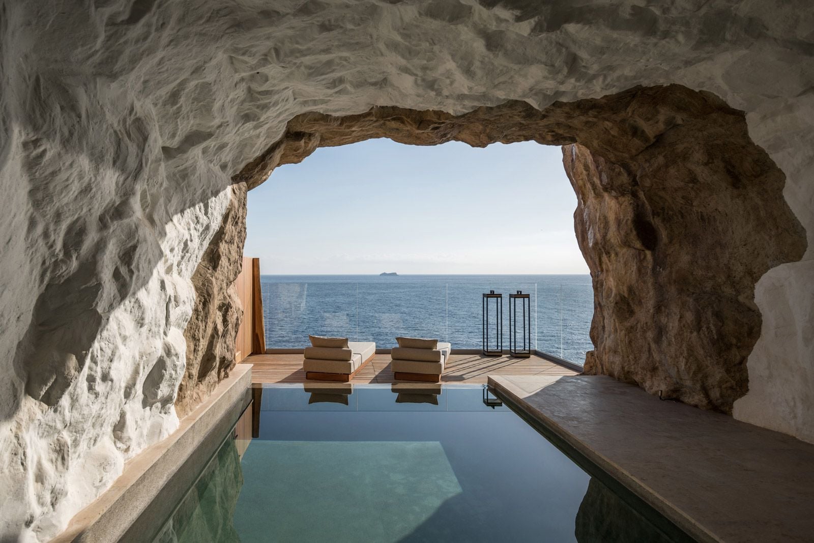 ACRO Suites, Europe — Limestone cave interior with infinity pool overlooking Mediterranean Sea, minimalist wooden loungers, natural stone architecture and coastal horizon.